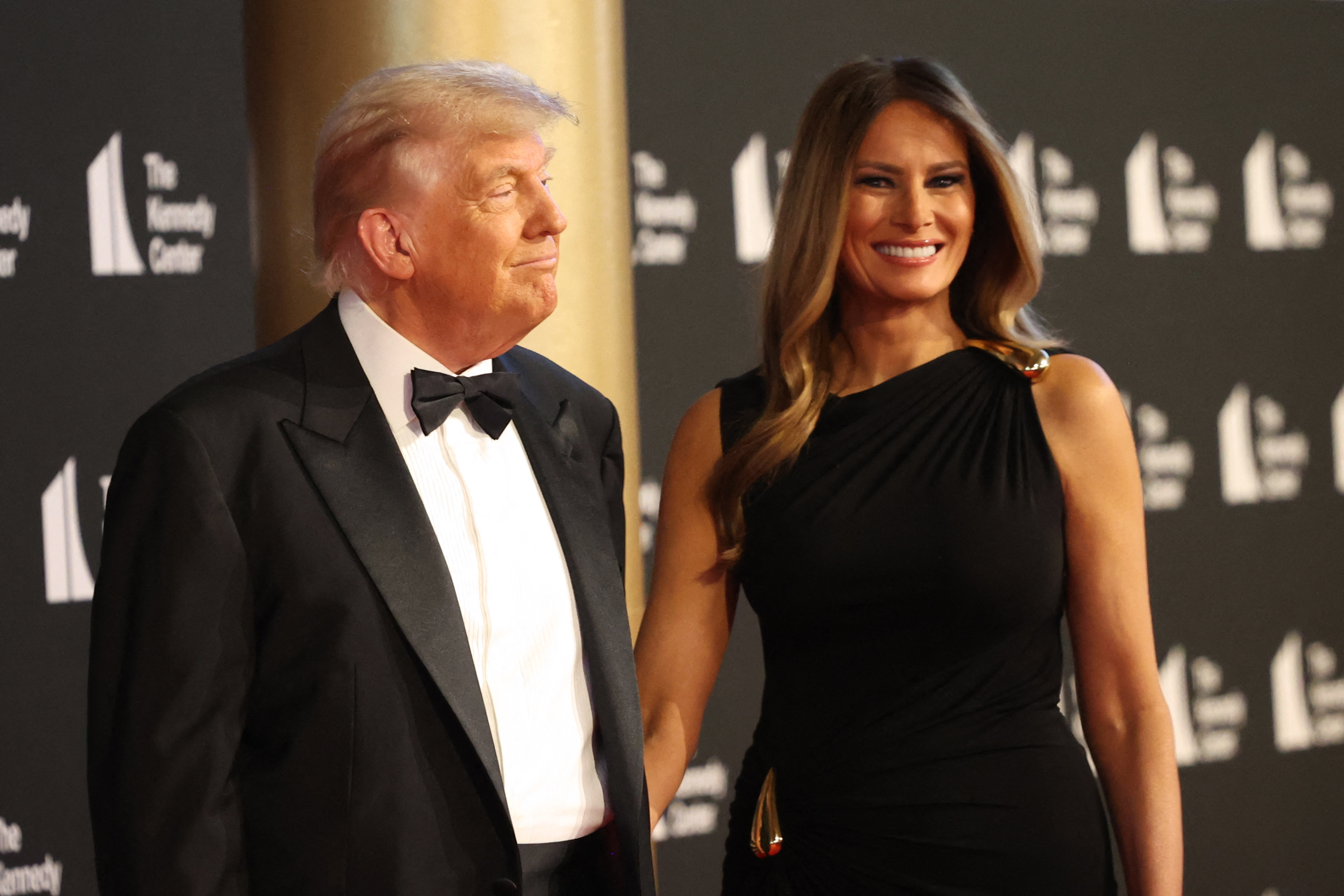 More of Melania’s dress is visible here, showing a ruched design at the waist with gold detailing that catches the light. She smiles brightly, adding warmth to the formal evening.