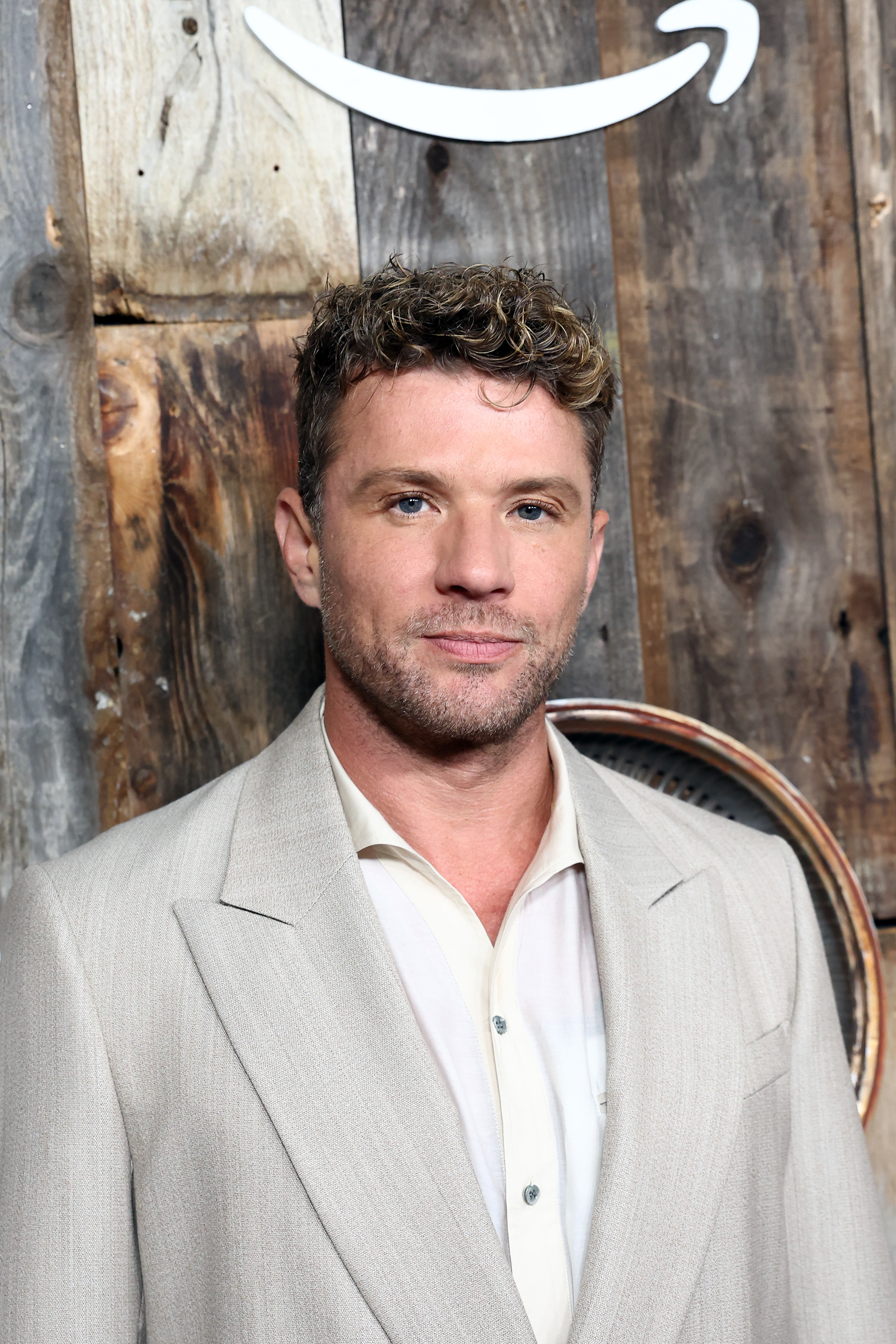 Ryan Phillippe poses solo at the "Motorheads" red carpet and screening held at the Academy Museum's Ted Mann Theater, dressed in a sand-toned suit over a crisp white shirt, exuding understated elegance against a rustic wooden backdrop.