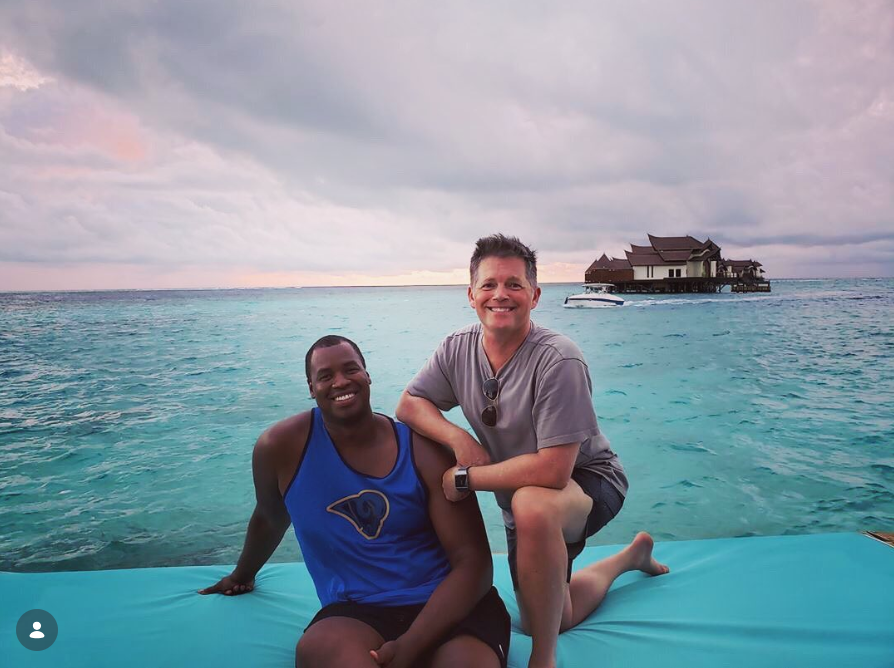 Jason Collins and his husband, Brunson Green, pose together on a floating dock surrounded by turquoise ocean water during a tropical getaway, with overwater villas visible in the distance beneath a cloudy evening sky. | Source: Instagram/jasoncollins_98