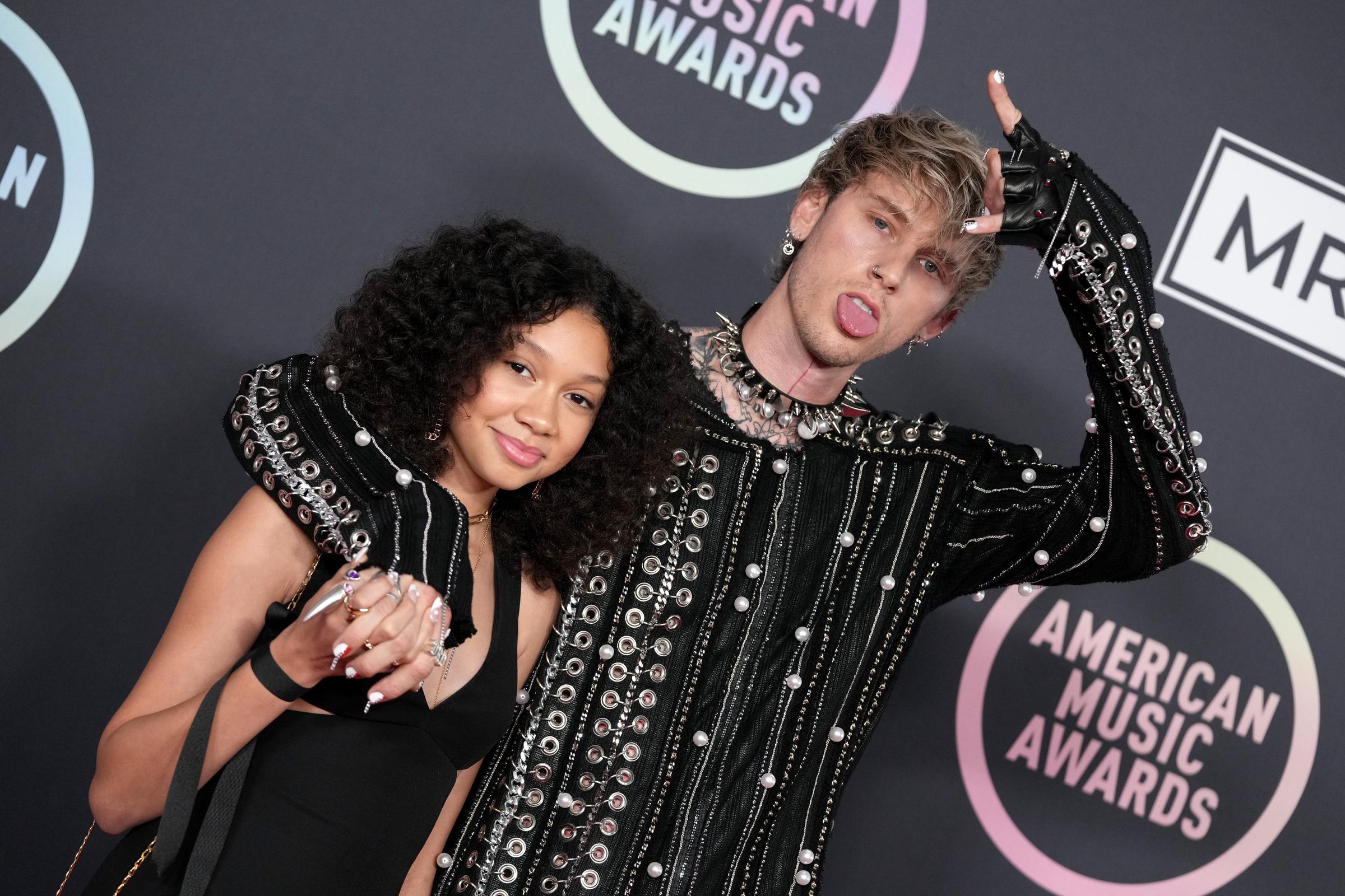 Casie Baker leans into Machine Gun Kelly's shoulder with a soft smile while he flashes a playful hand sign on the 2021 American Music Awards red carpet. The duo sport bold, black studded ensembles.