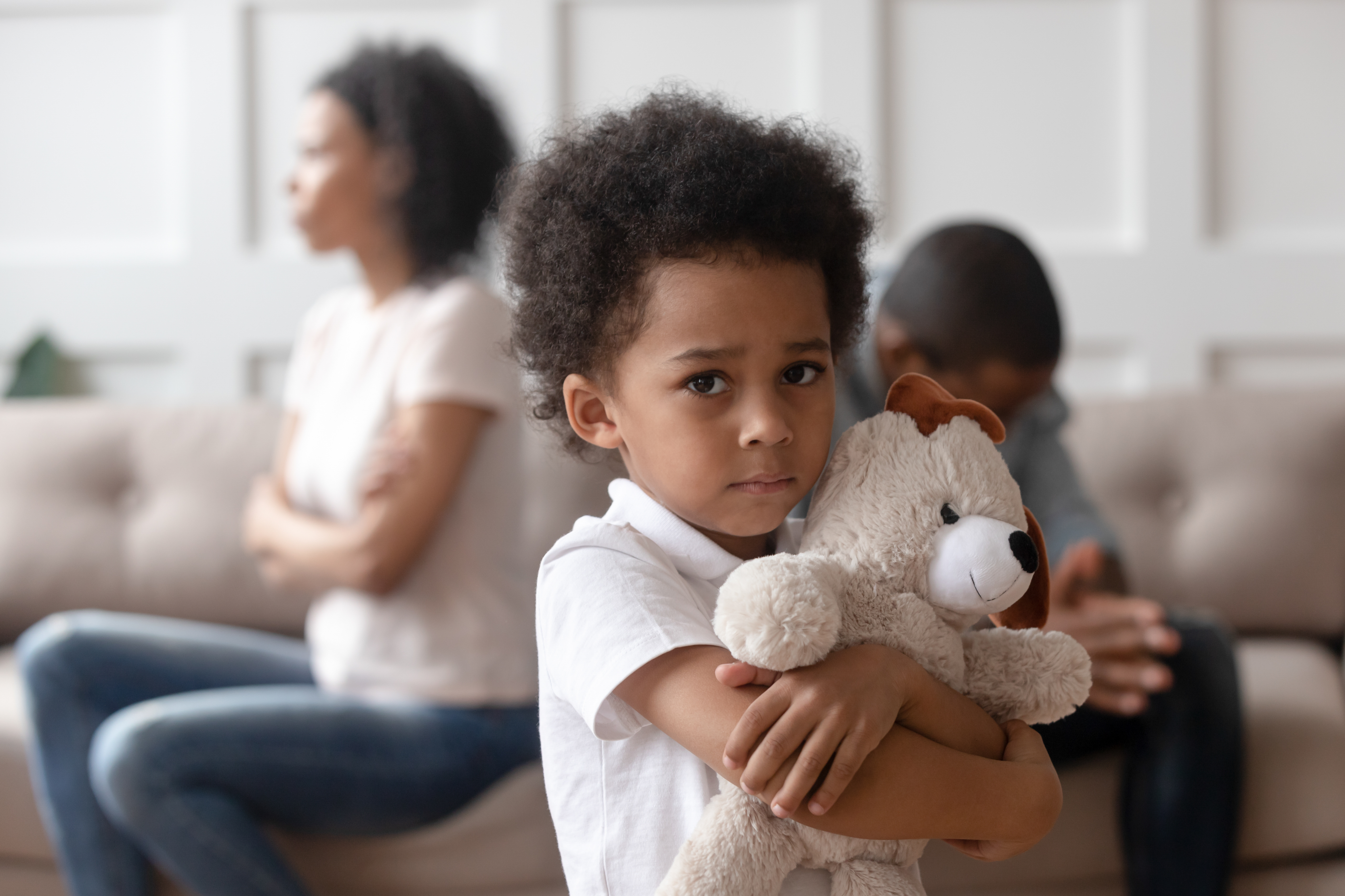 A sad Black child holding a teddy bear while a man and a woman turn their backs on each other in the background.  | Source: Shutterstock