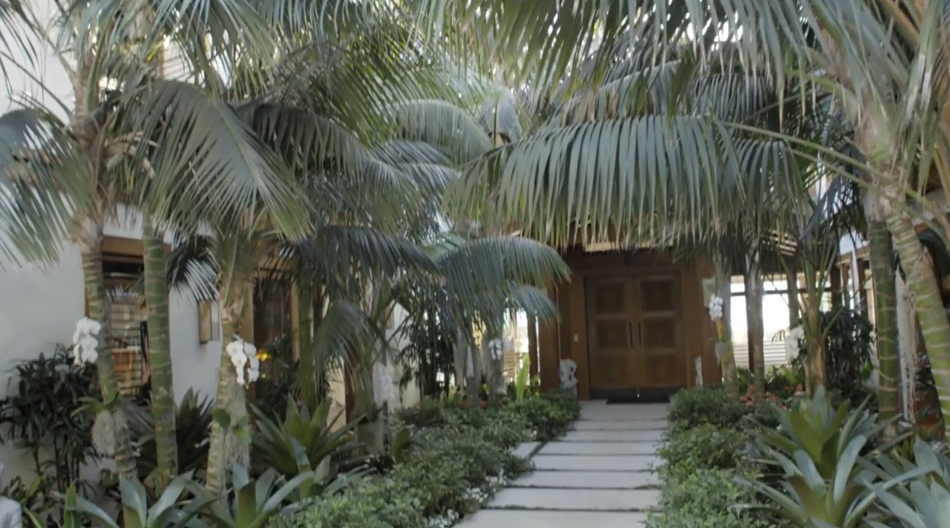 A stone-paved walkway winds through a lush courtyard filled with leafy tropical foliage and potted white orchids leading toward the grand teak entrance doors. Sunlight filters through the dense canopy of mature palm trees to highlight the home's warm wood paneling and natural stone accents. | Source: YouTube/Architectural Digest