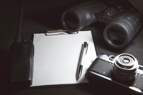 Blank page with copy space, pen, portable radio, binoculars and photo camera on a black spy agent table background. | Source: Shutterstock.
