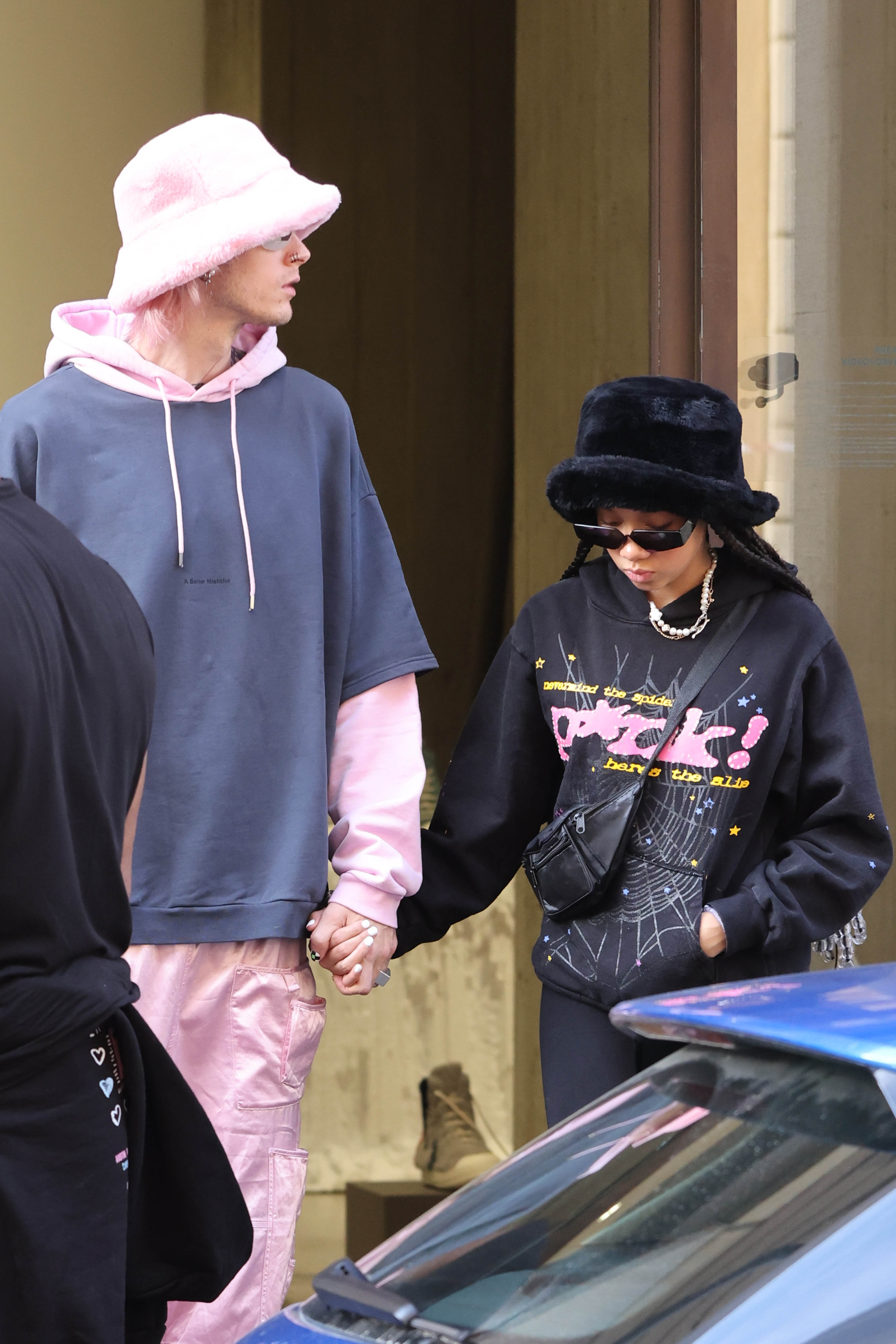 Machine Gun Kelly and Casie Baker hold hands while walking out of a Milan doorway in coordinated streetwear. Casie looks down in dark sunglasses and a fuzzy bucket hat, while MGK glances left.