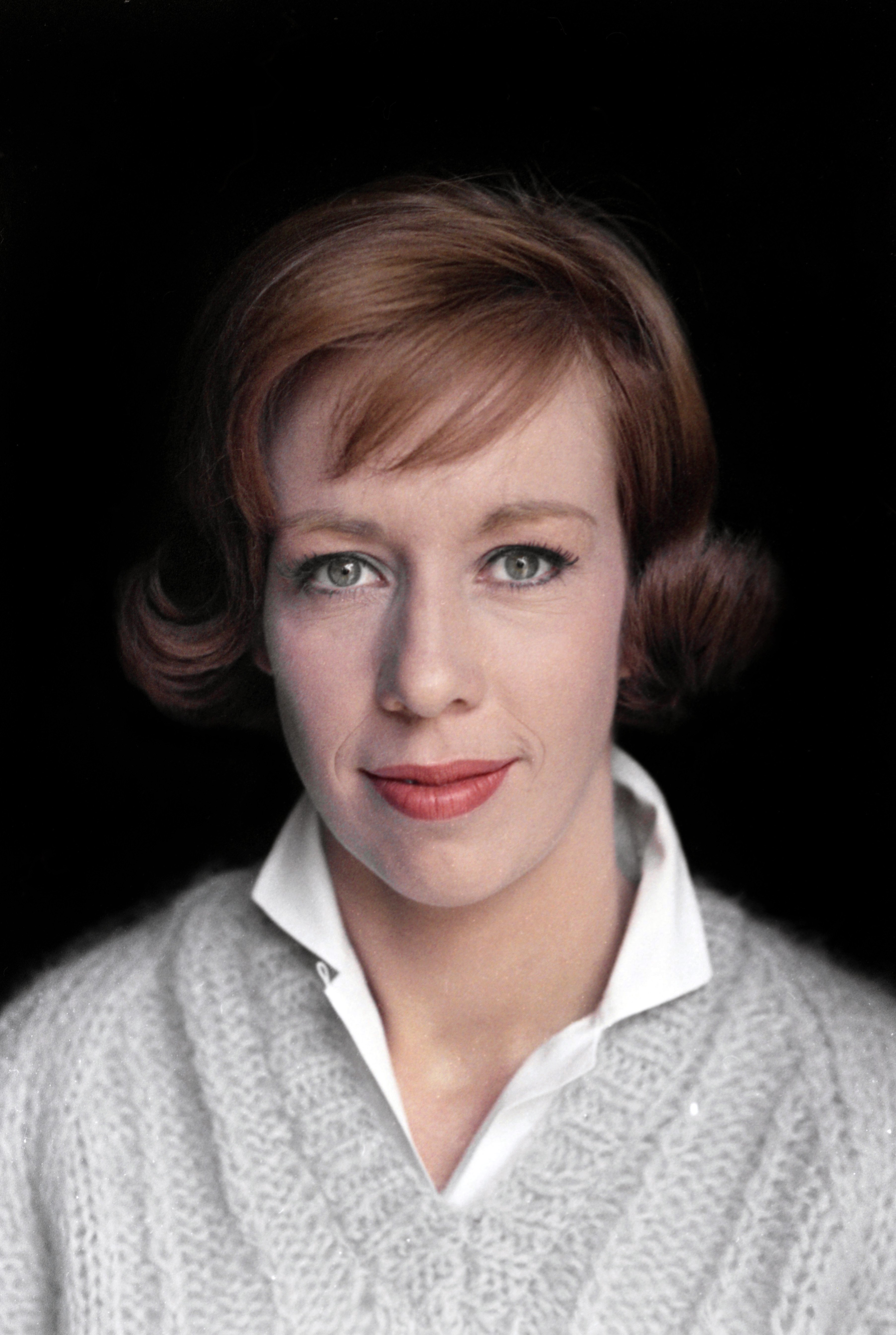 In a studio portrait, she wears a collared white shirt under a textured knit sweater. The shot is tightly framed on her face, with her hair styled neatly and her gaze directed straight at the camera.