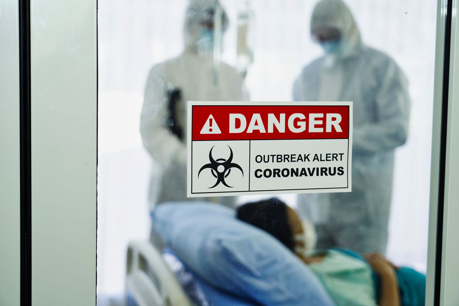 A coronavirus danger sign in front of a control area with a team of doctors in protective gear attending to an infected patient in a quarantined room | Photo: Shutterstock/Kobkit Chamchod