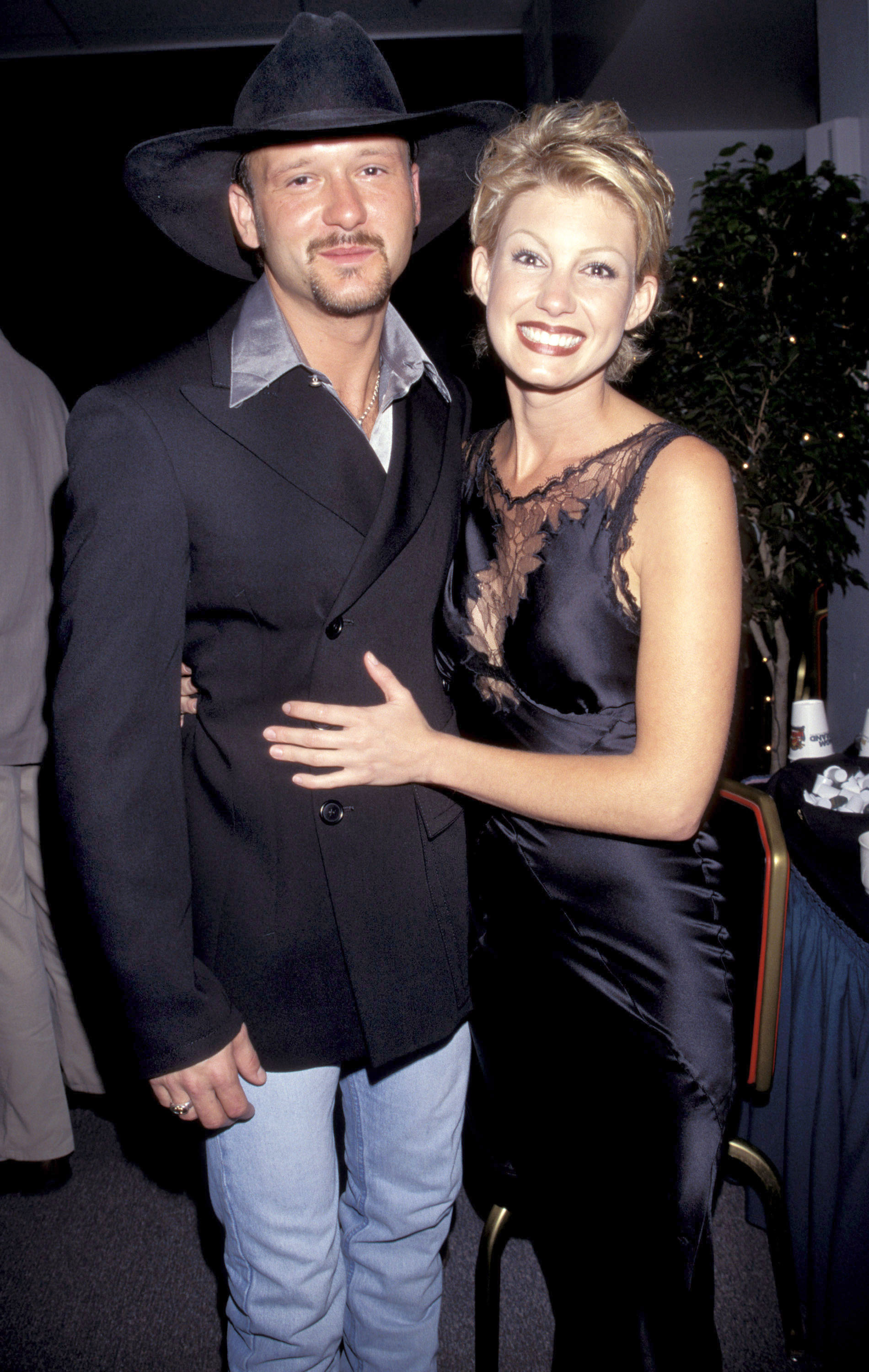 Faith Hill wraps her arm around Tim McGraw in a playful, close pose as they smile for the camera. A warm and early moment between the country stars, full of charm and old-school glamour.