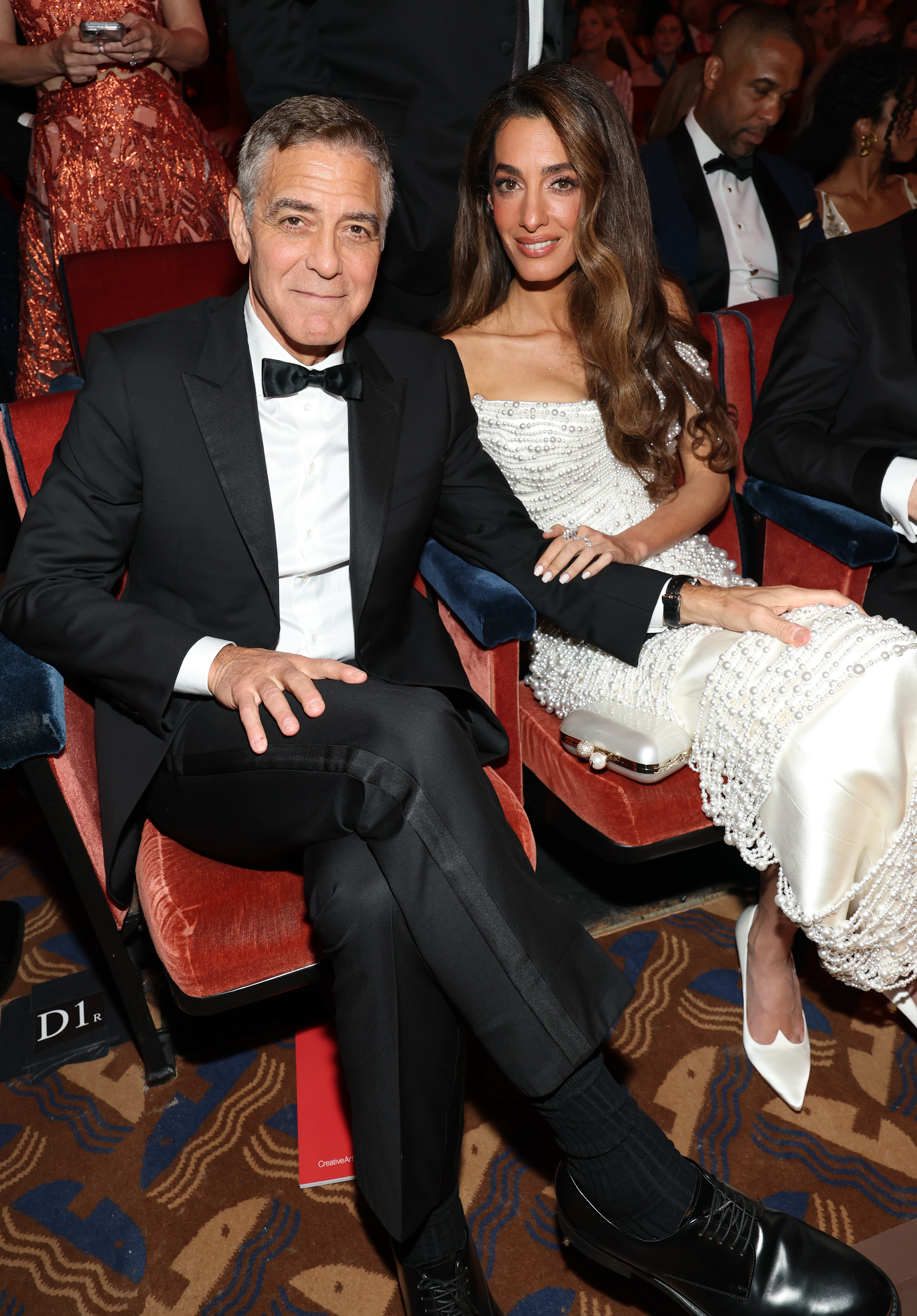 Seated inside the venue, George Clooney sits with legs crossed while Amal leans toward him, one hand resting on his arm. The couple strikes a relaxed pose, surrounded by other guests as they await the start of the ceremony.