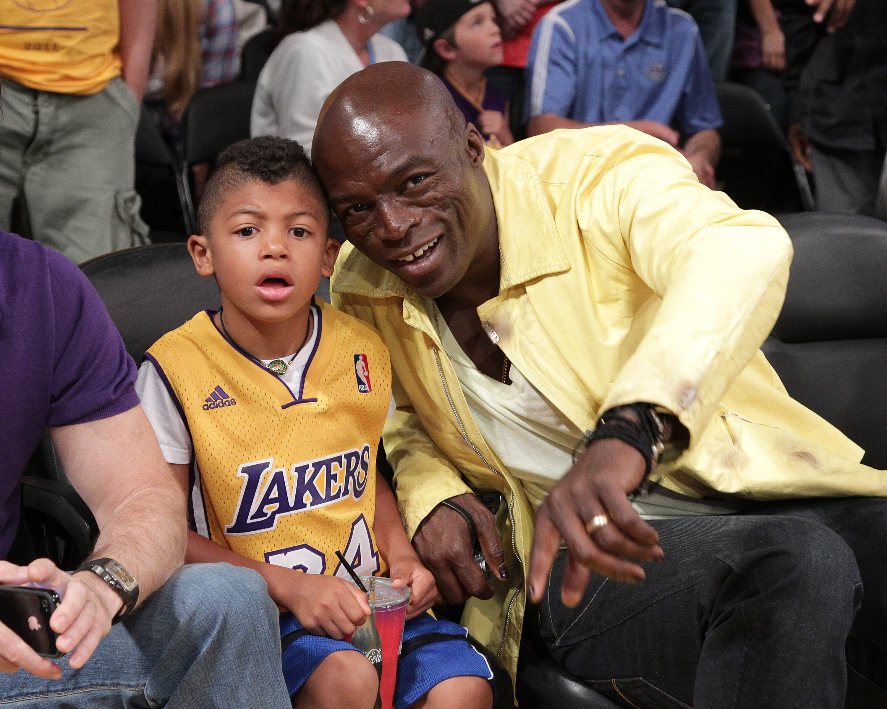 Leaning in with a bright, easy smile, Seal shares a lively courtside moment with Henry Samuel, who watches the action with wide-eyed focus at an NBA game in Los Angeles.