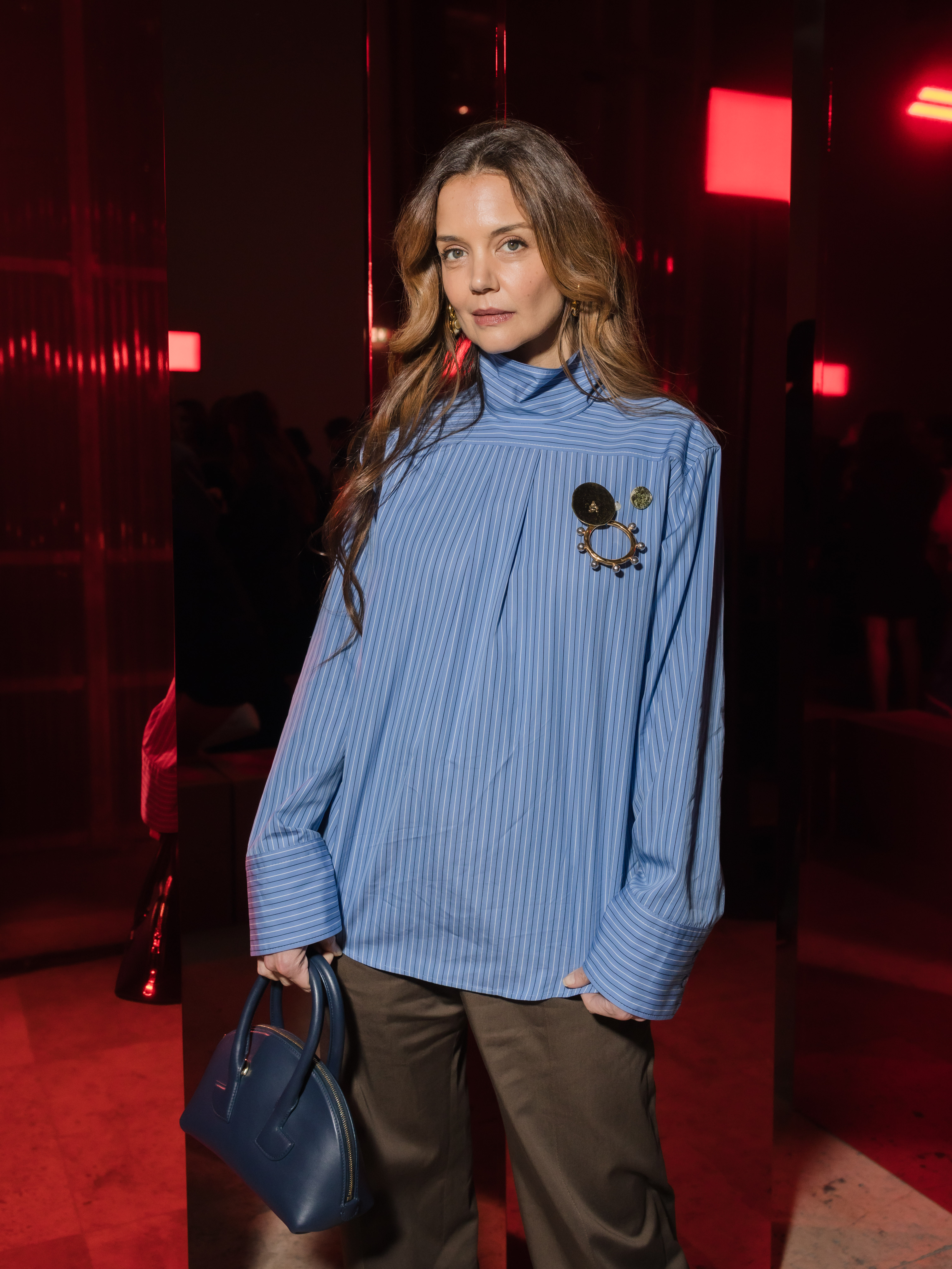 Katie Holmes embraced avant-garde fashion energy in an oversized blue striped top styled with flowing trousers and a structured handbag. The dramatic red-lit venue added to the effortlessly cool and artistic mood of her Paris Fashion Week appearance.