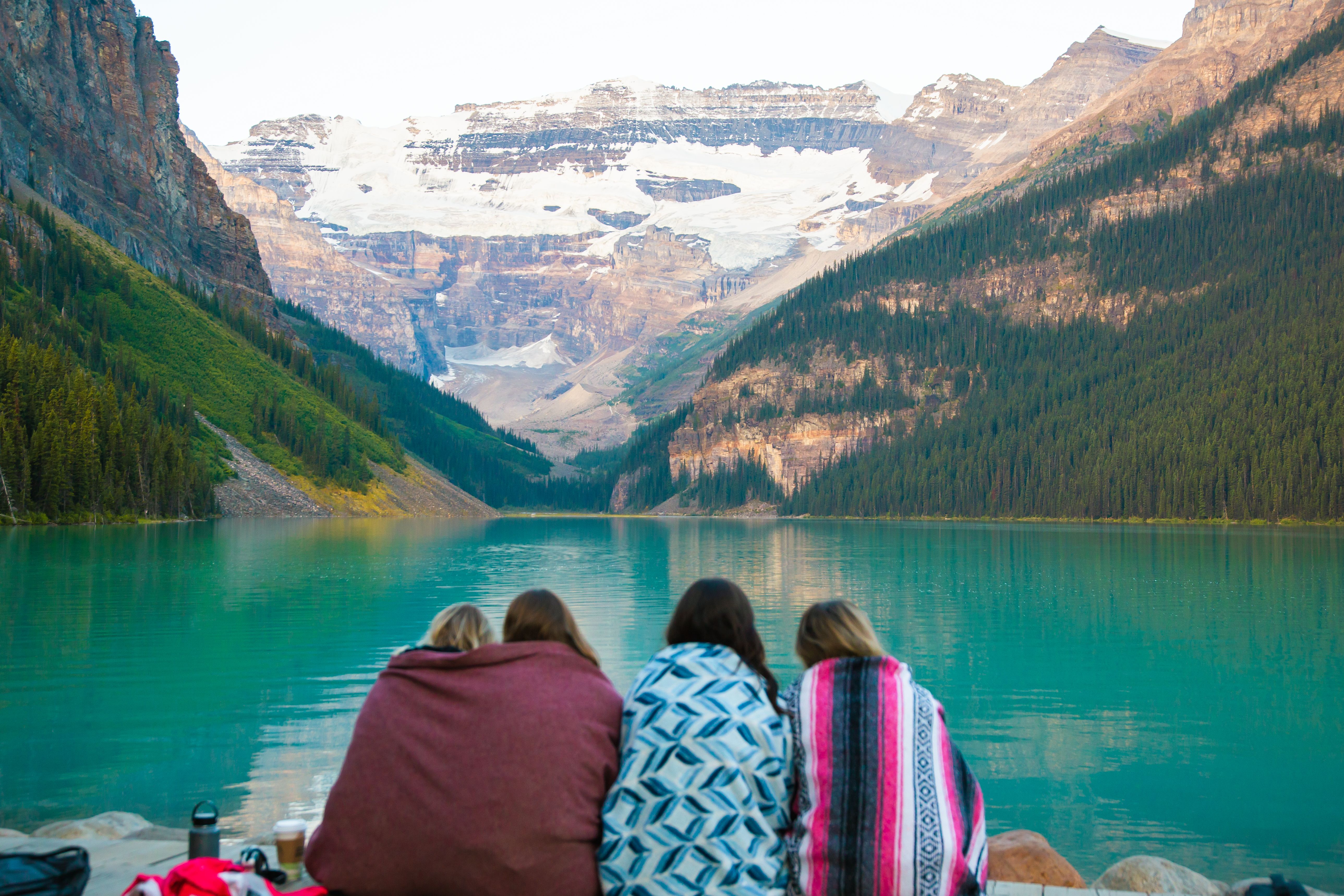 Four women wrapped in blankets sitting by a turquoise glacial lake, facing snow-capped mountains | Source: Pexels