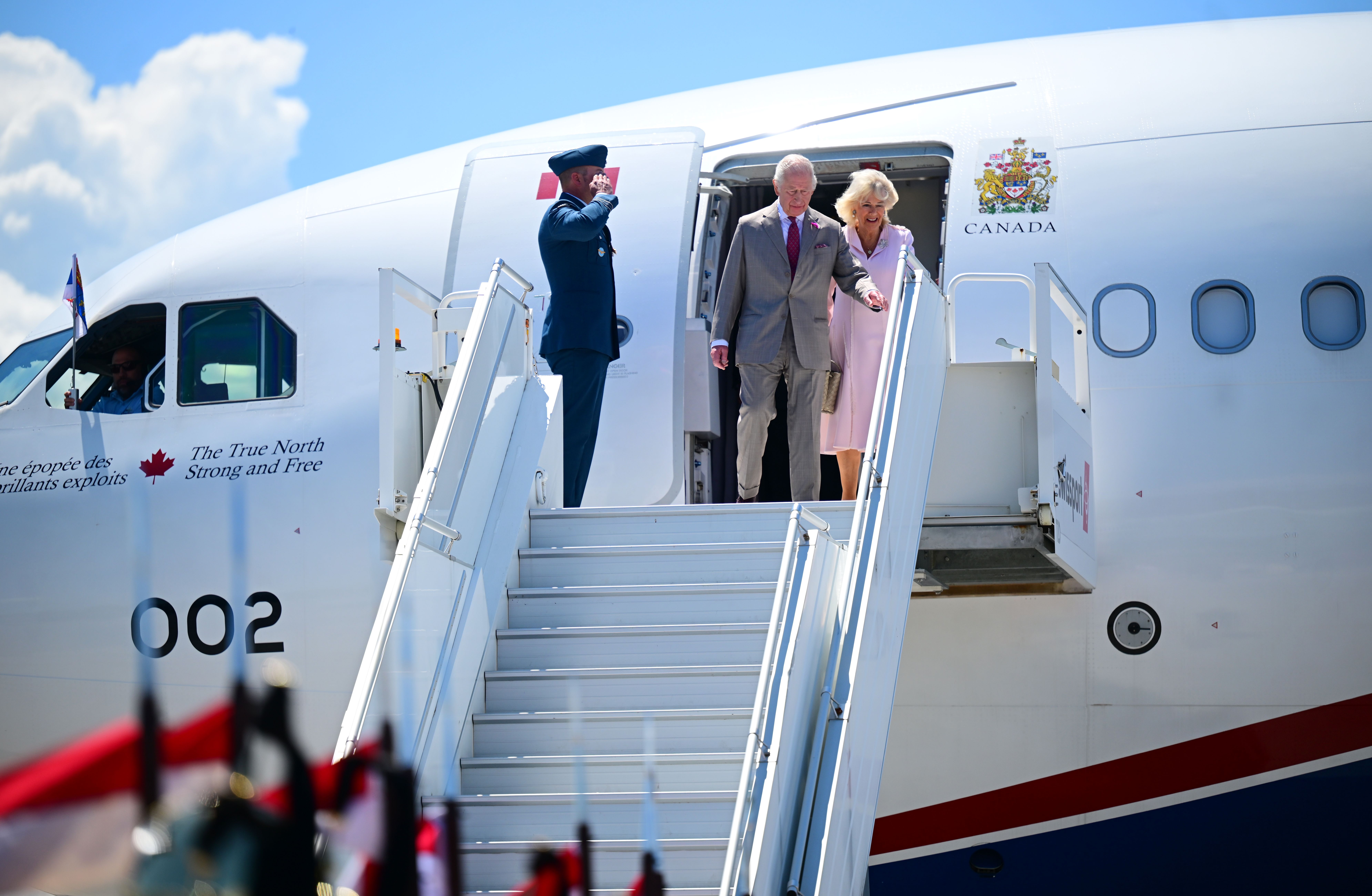 King Charles III and Queen Camilla stand at the door of the plane beside a saluting officer, preparing to descend the stairs upon arrival at Ottawa Airport during their official visit to Canada.