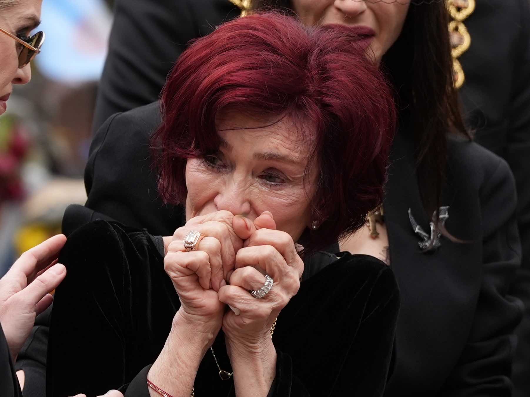 Ozzy's widow, Sharon clutches her hands to her face, visibly overcome with grief during Ozzy Osbourne's funeral. Her tear-filled eyes and trembling expression capturing the raw pain of saying goodbye to her beloved husband.