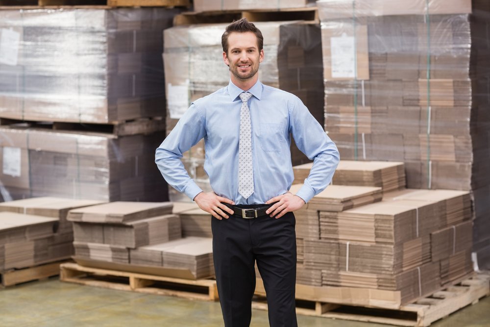 A smiling manager placing his hands on his hips in a large warehouse. | Photo: Shutterstock.