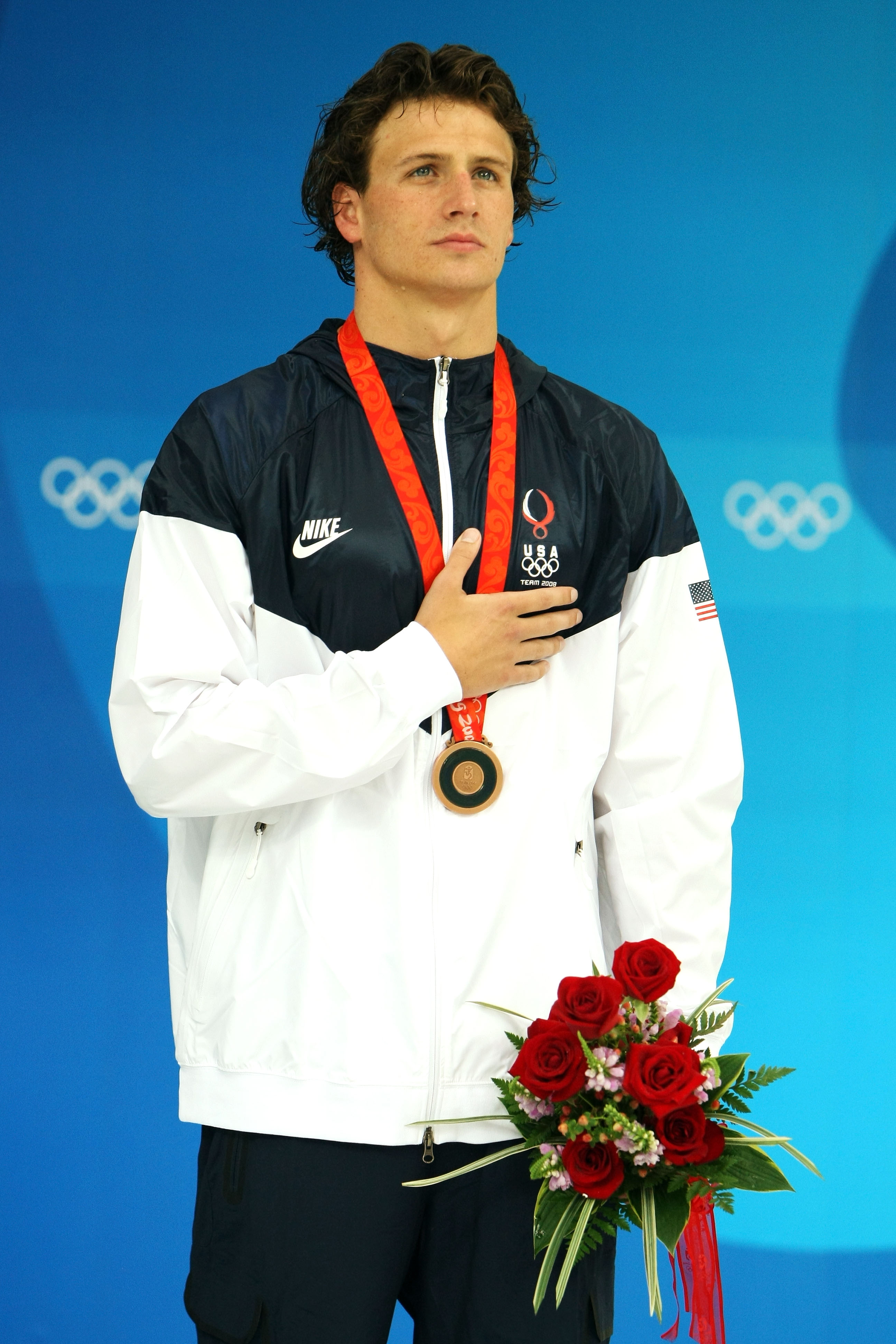 Ryan Lochte stands solemnly on the Olympic podium with a bronze medal draped around his neck, his hand pressed against his chest during the ceremony. He also holds a bouquet of red roses while wearing a Team USA jacket at the National Aquatics Center.