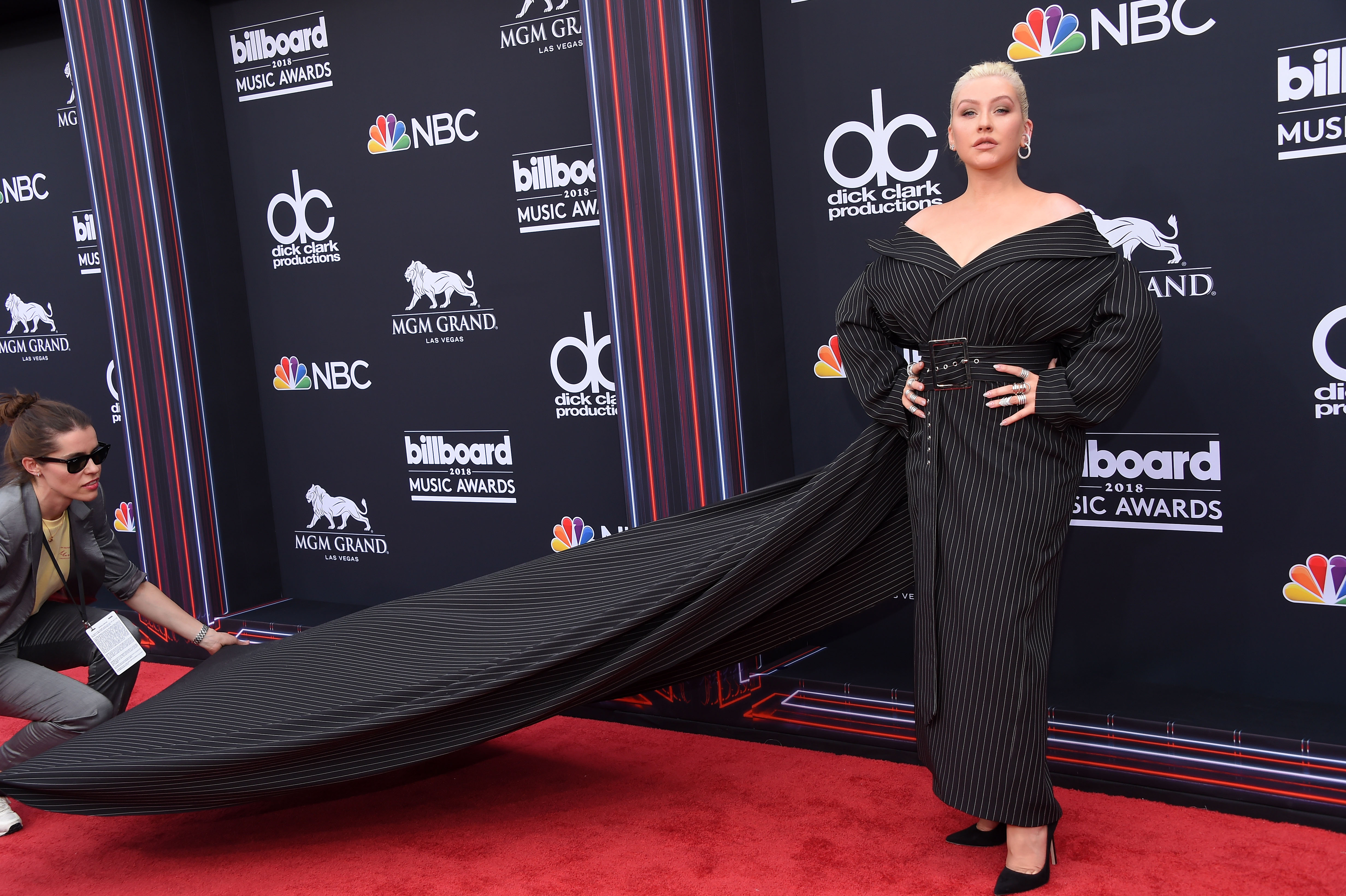 Christina Aguilera makes a dramatic entrance at the 2018 Billboard Music Awards at the MGM Grand in Las Vegas. She wears an oversized, pinstriped gown with a structured off-the-shoulder neckline, cinched waist, and extended train — a bold fashion moment that showcased her shift toward more avant-garde red carpet looks.