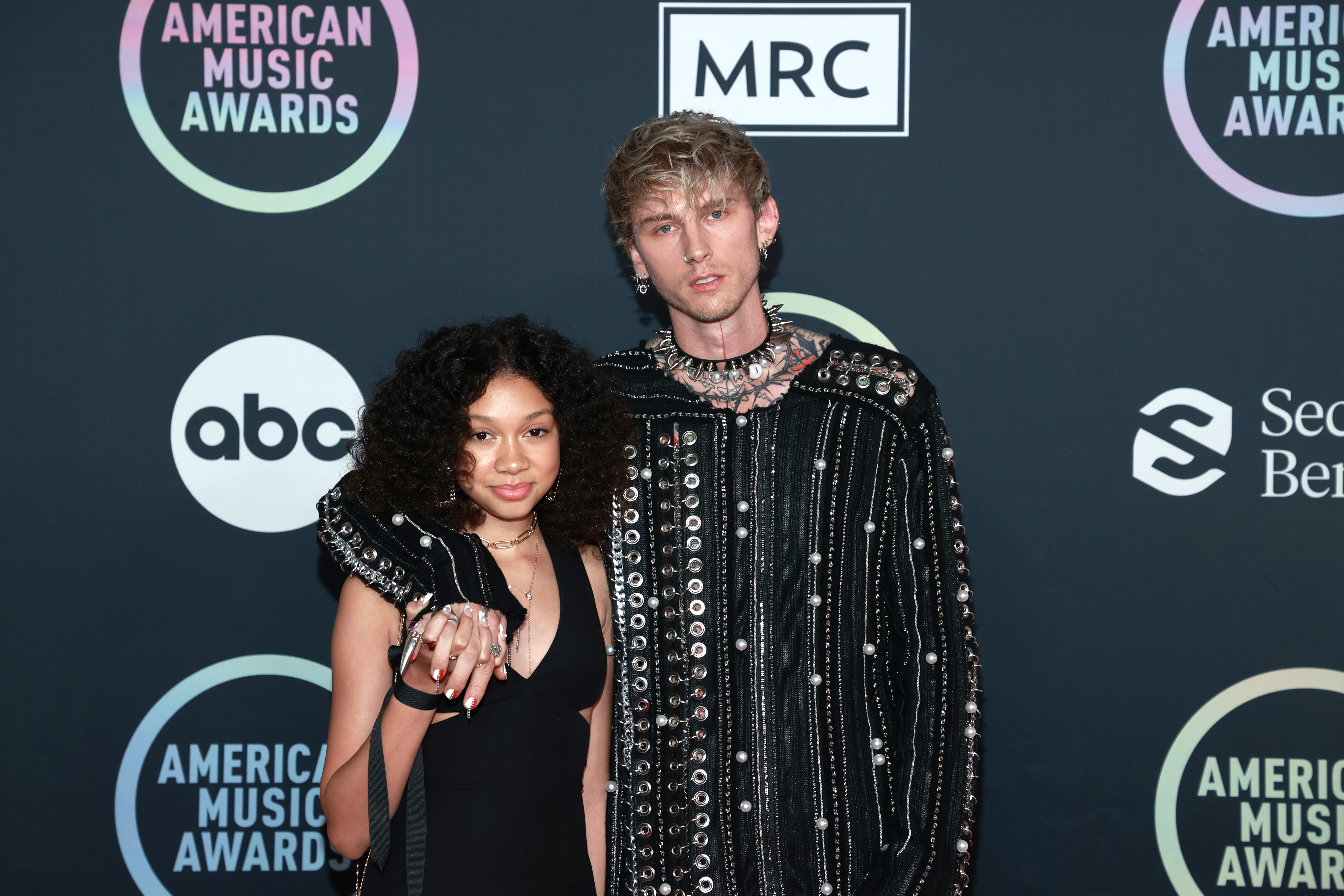 Standing close with fingers interlocked, Casie Baker and Machine Gun Kelly pose head-on at the 2021 AMAs. Their serious expressions match their coordinated gothic looks.