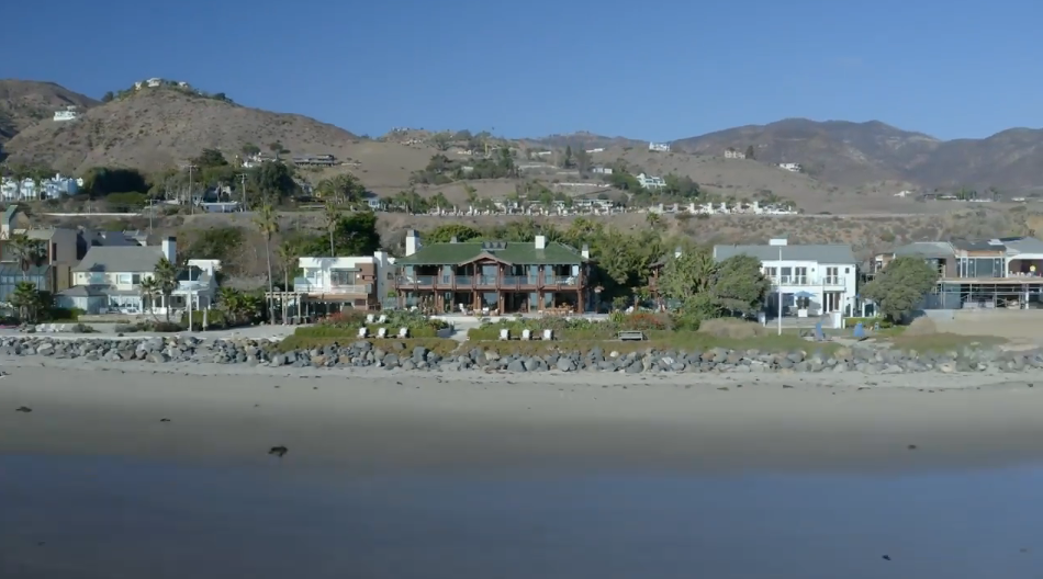 A wide aerial view shows the beachfront compound situated along the coastline, set against a backdrop of rolling Malibu hills. Rock revetments line the sandy shore in front of the home, shielding the property while offering uninhibited views across the ocean. | Source: YouTube/Architectural Digest