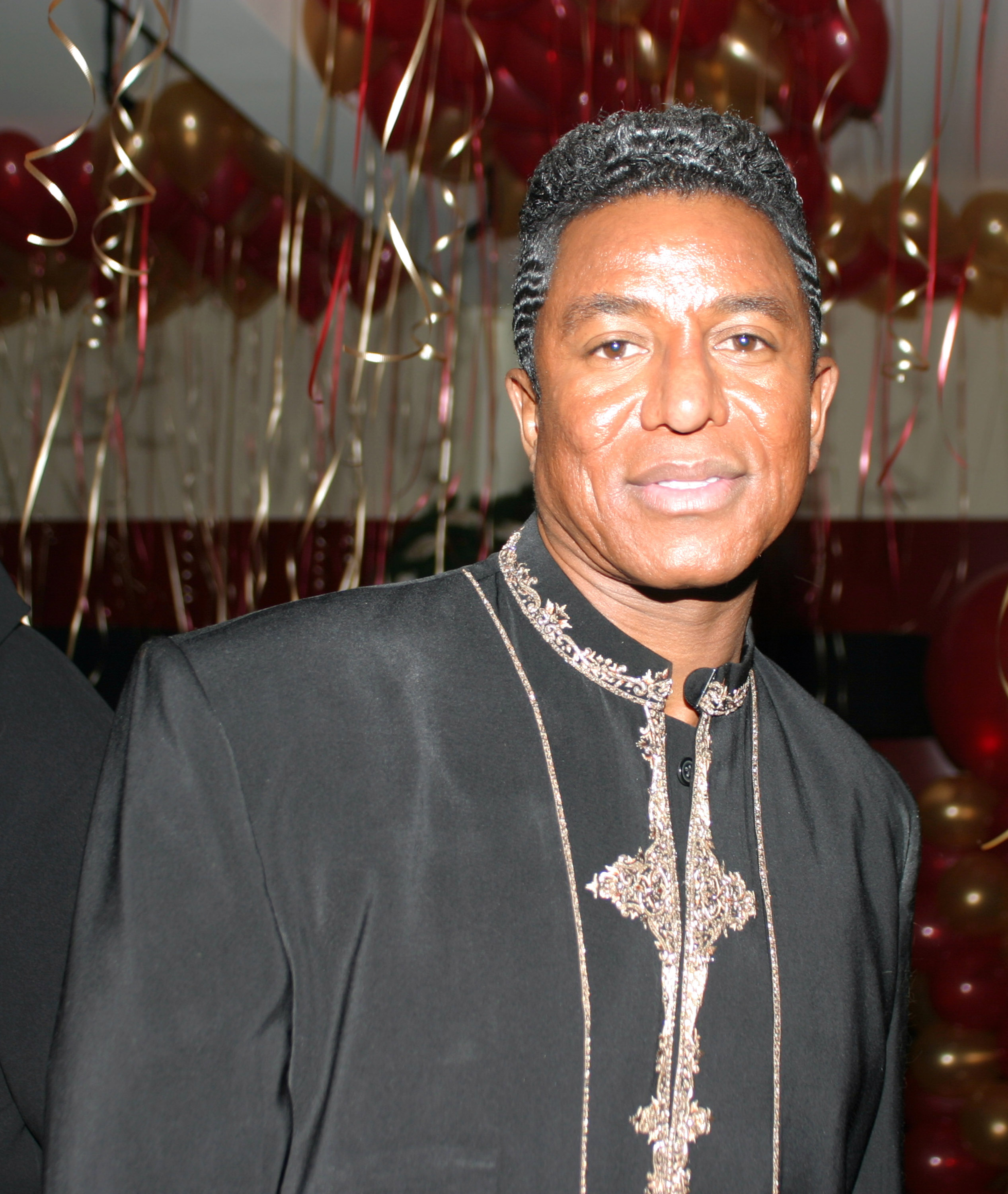 Jermaine Jackson poses at Kim Porter’s birthday celebration, his embroidered black jacket standing out against a festive backdrop of red and gold balloons. The intricate detailing and polished finish elevate the look, matching the upscale party atmosphere.