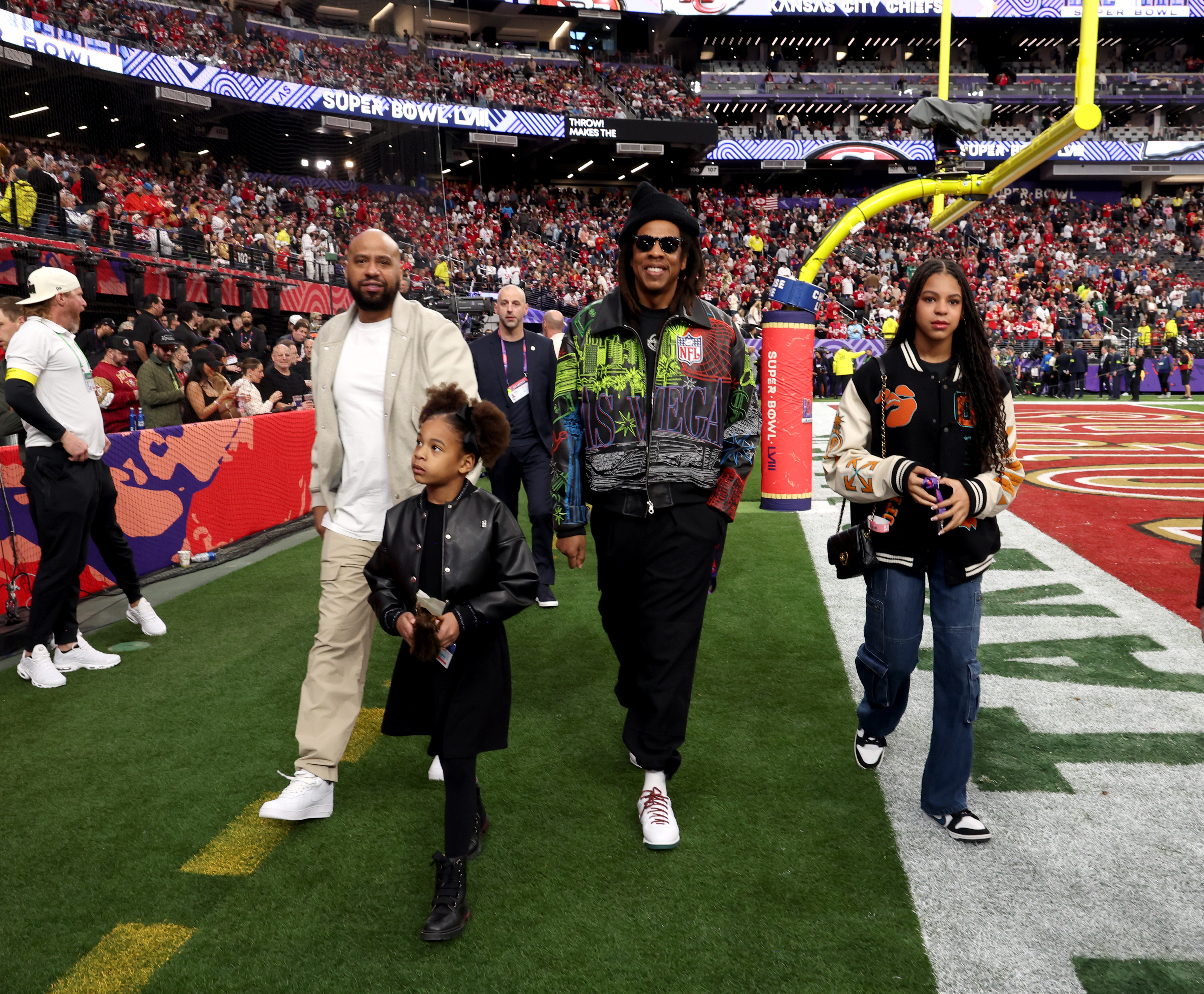 Jay-Z moves down the field with Blue Ivy and Rumi close by, framed by stadium lights and fans in the stands. The image captures the family in motion as the venue fills.