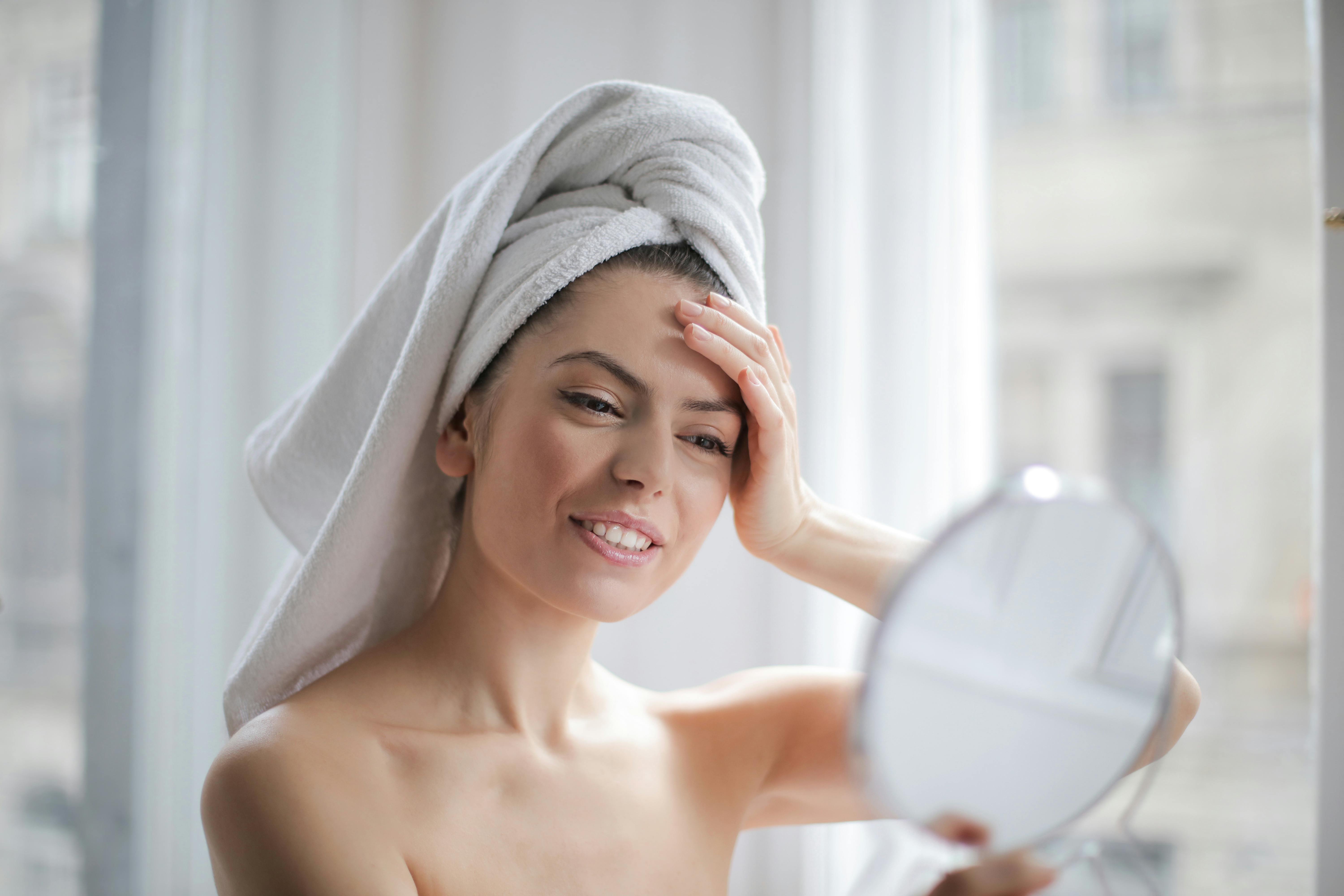 A happy woman looking at herself in the mirror while wearing a towel to dry her hair | Source: Pexels