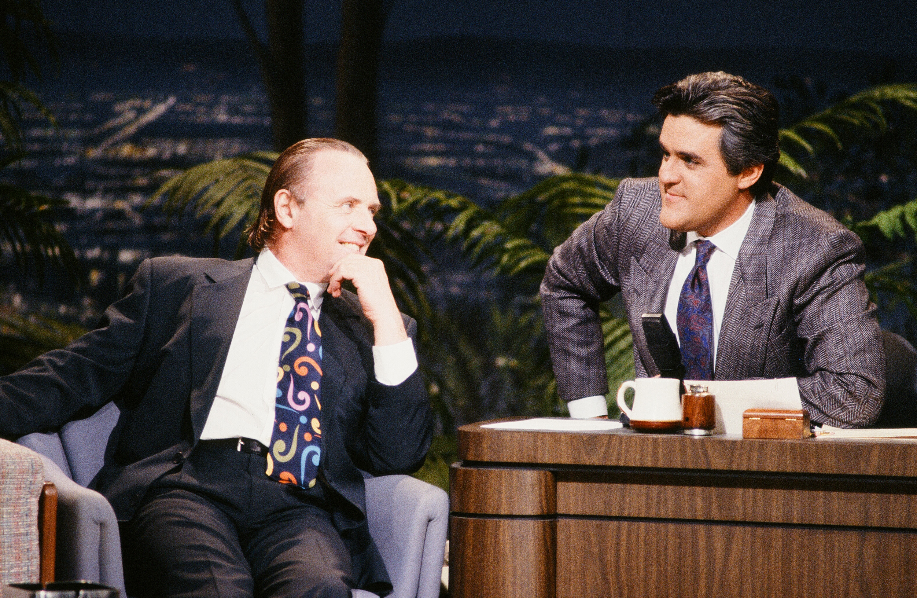 Anthony Hopkins shares an engaging moment with Jay Leno during an interview, both men animated with laughter and ease. Hopkins' colorful tie adds a playful flair to his sleek suit, reflecting his charismatic energy. Under the studio lights, their lively exchange captures the warmth and wit of late-night television at its finest.