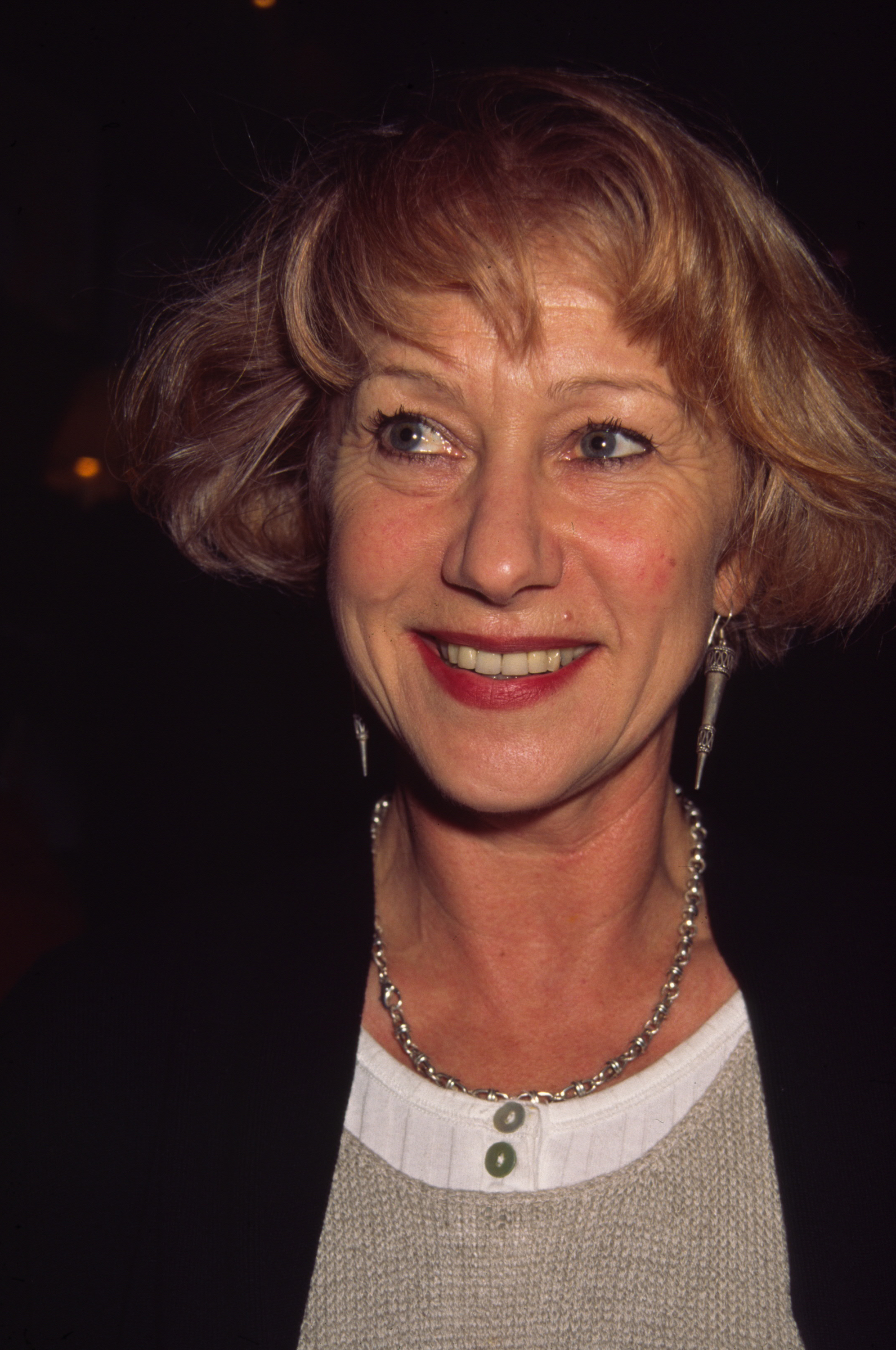 Helen Mirren beams in a candid close-up, her short tousled hair softly framing her face as she pairs silver drop earrings with a chunky chain necklace and a layered knit ensemble, exuding warmth and understated charm.