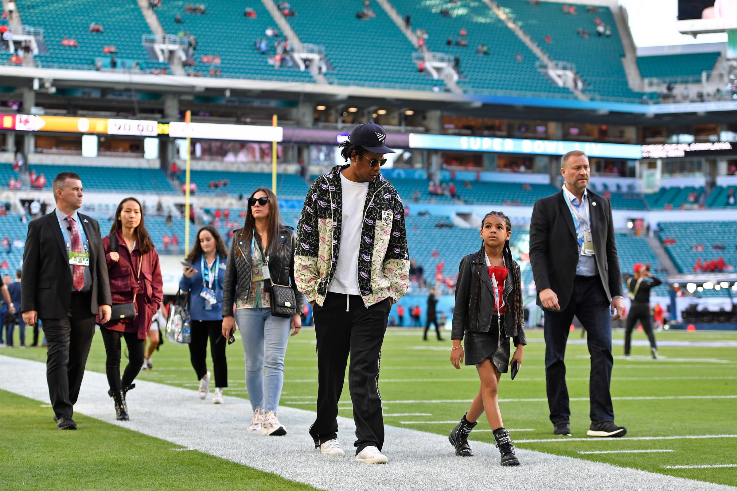 Jay-Z walks the field alongside Blue Ivy during pregame, flanked by staff as they take in the stadium before kickoff. The moment captures a relaxed walk-through ahead of the game.