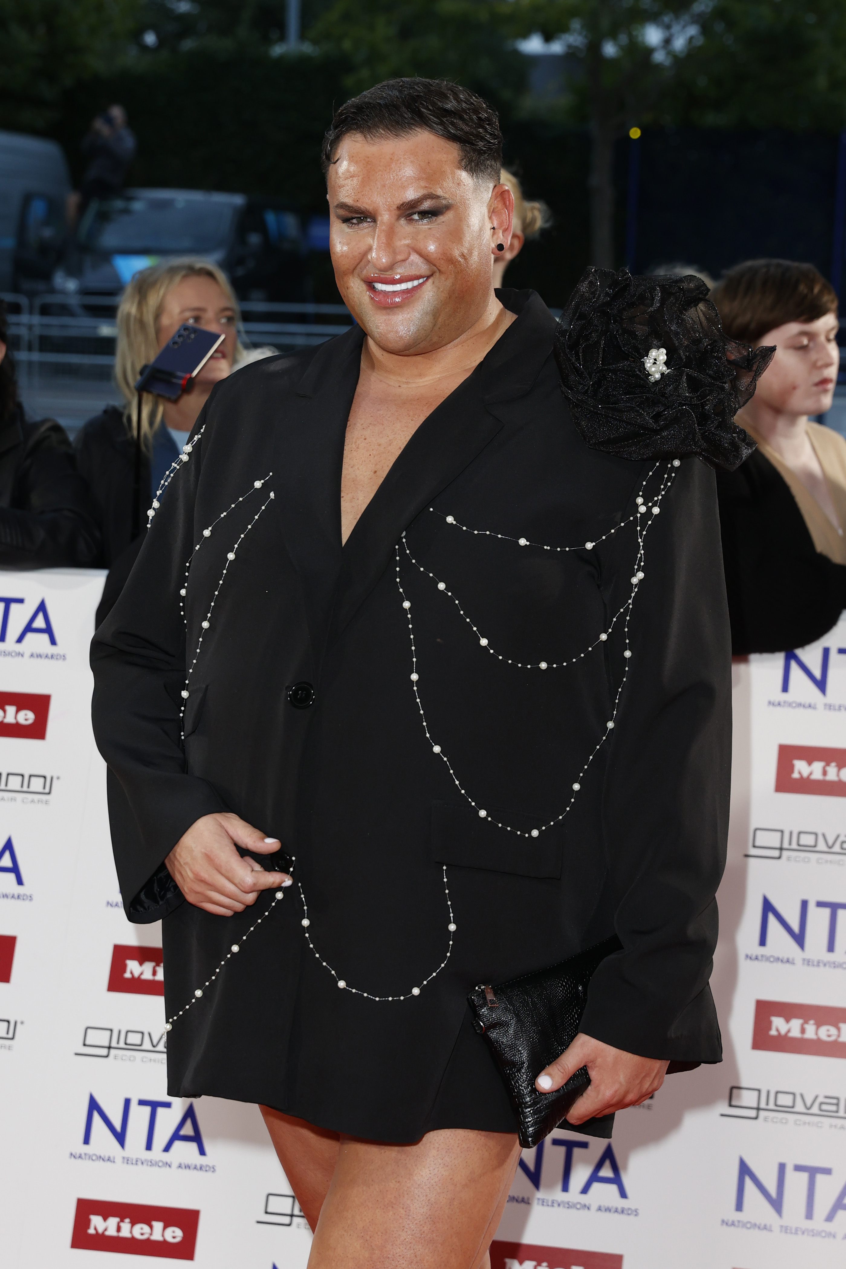 Potts stuns in statement couture at the 2025 National Television Awards, arriving at The O2 Arena on 10 September in a black blazer dress adorned with pearl chain draping and a dramatic floral shoulder detail. With a sleek black clutch in hand and a glossy, confident smile, the reality star owns the spotlight once again.