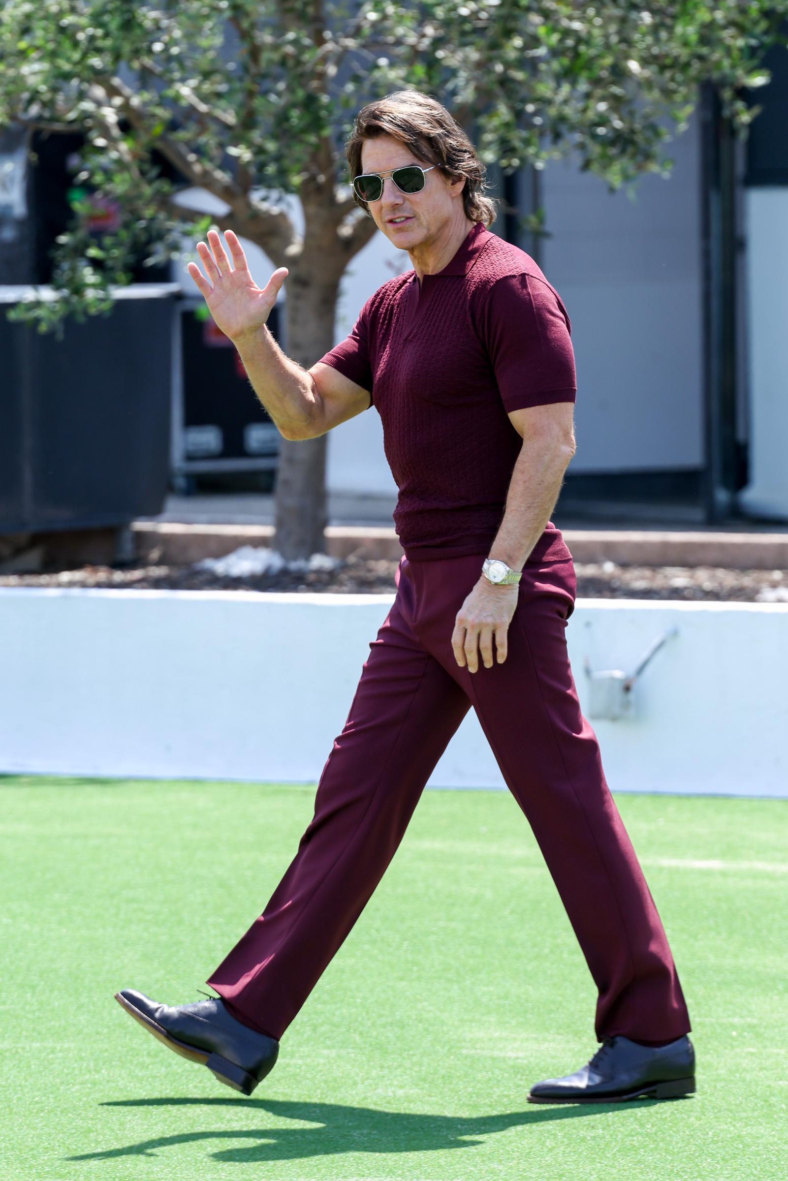 Tom Cruise walks across a grassy terrace outside the Palais des Festivals, offering a wave to fans and photographers. He kept his look sleek and coordinated in burgundy during the "Mission: Impossible – The Final Reckoning" photocall.