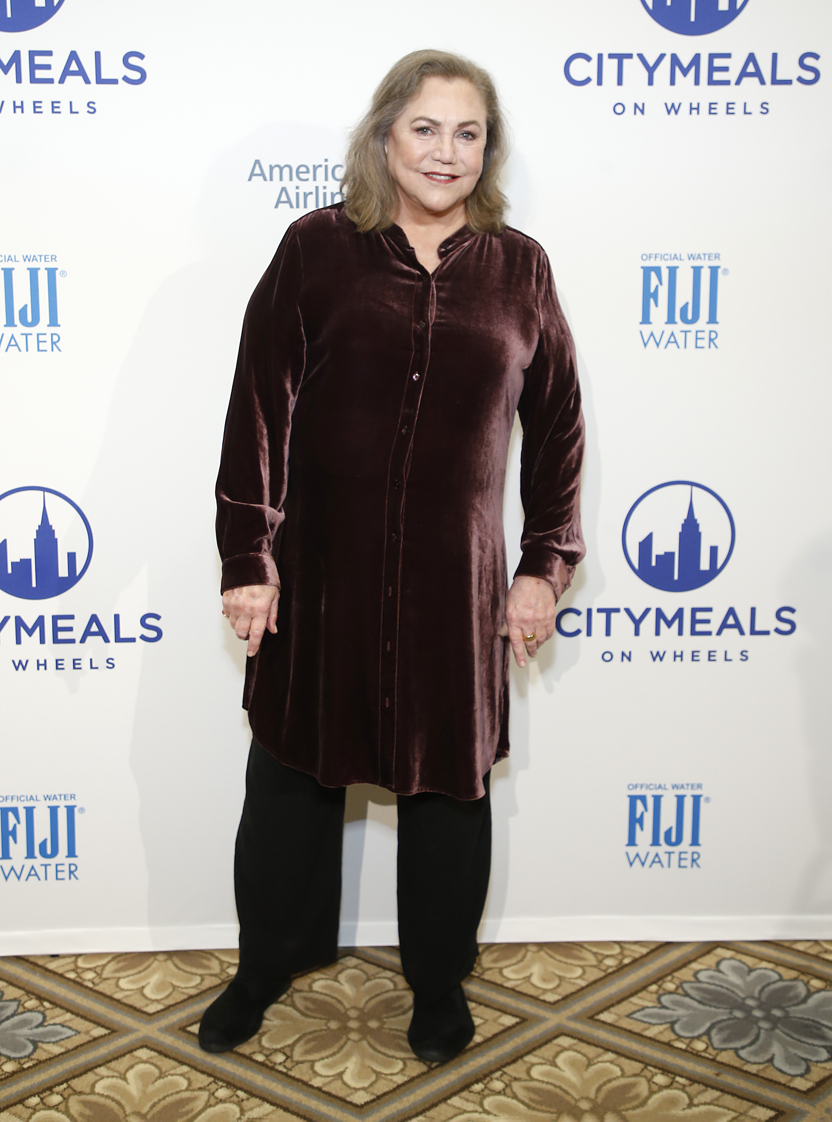 Kathleen Turner is seen at the Citymeals On Wheels' 34th Annual Power Lunch at The Plaza Hotel in New York City. She wears a rich burgundy velvet button-down tunic paired with black pants and shoes, giving her a polished yet comfortable look for the charity event.