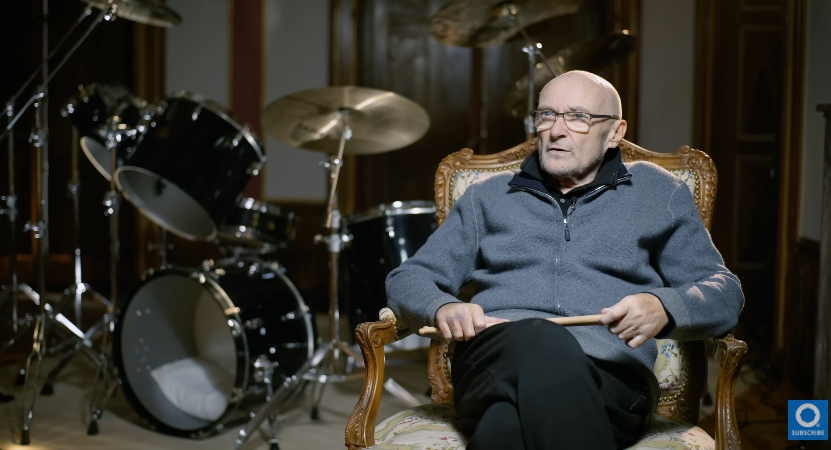 Though no longer able to play as he once did, Collins holds a pair of drumsticks in this rare portrait beside a full drum kit — a poignant visual reminder of the instrument that shaped his legacy. Now requiring assistance to walk, the image contrasts his physical limitations with the powerful musical identity he spent decades building. | Source: YouTube/Drumeo