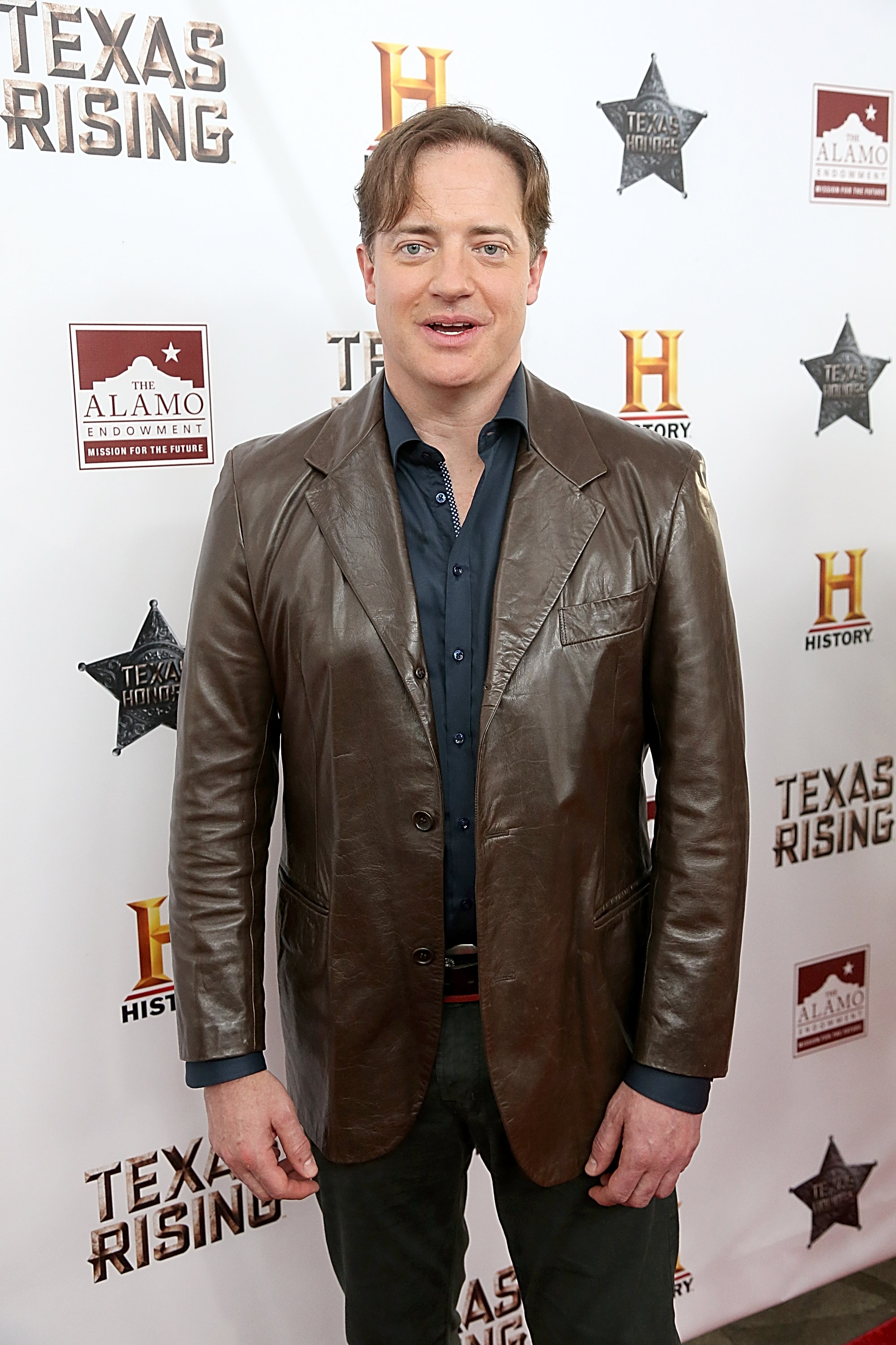 Brendan Fraser smiles in a brown leather jacket and dark shirt at the premiere of History's miniseries "Texas Rising," held at The Alamo in San Antonio, Texas.