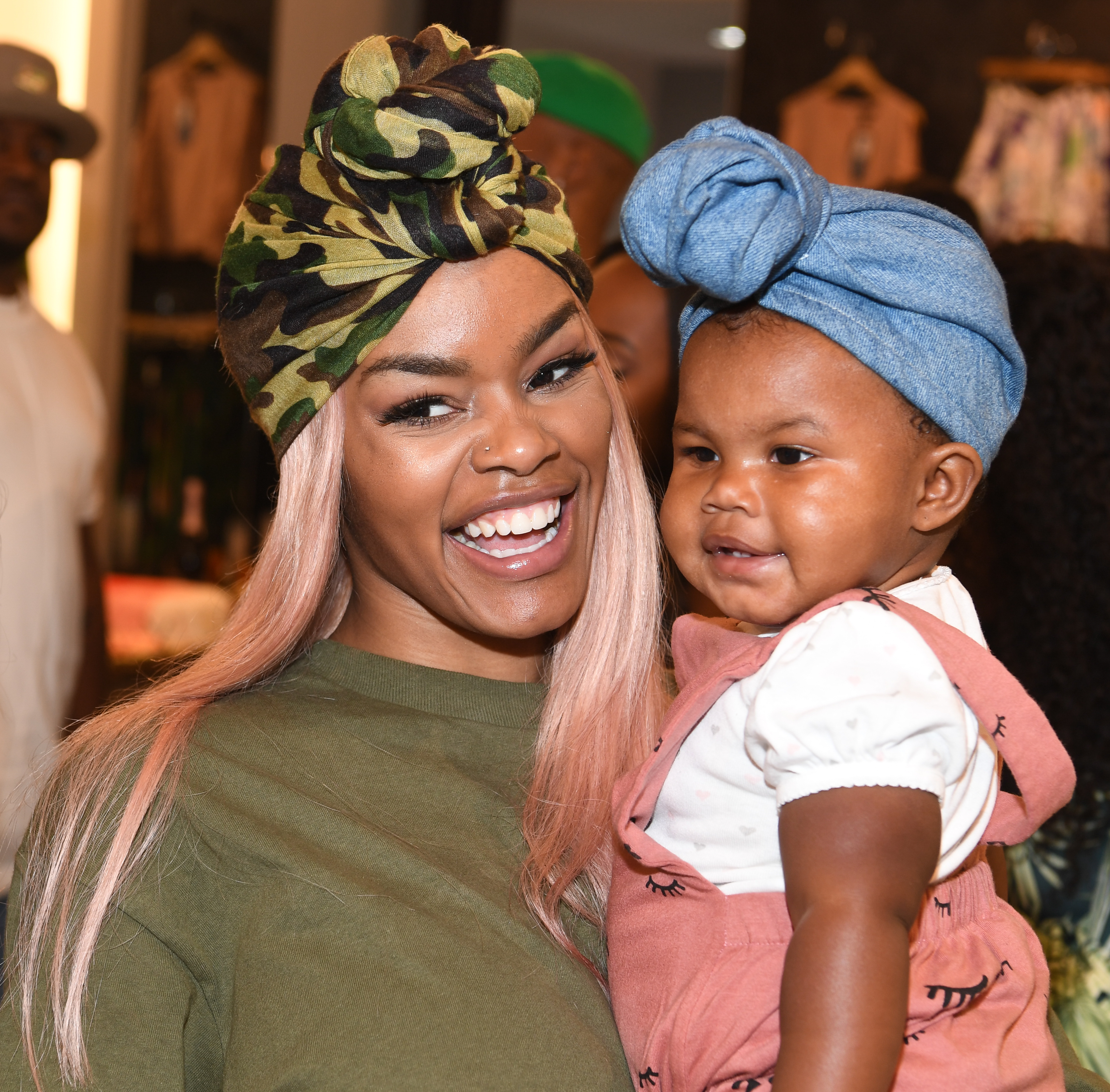 Teyana Taylor beams while holding Iman Tayla "Junie" Shumpert Jr. at a Baby Buddha Bug Collection event in Atlanta. Mother and daughter wear stylish head wraps in the warm, joyful snapshot.