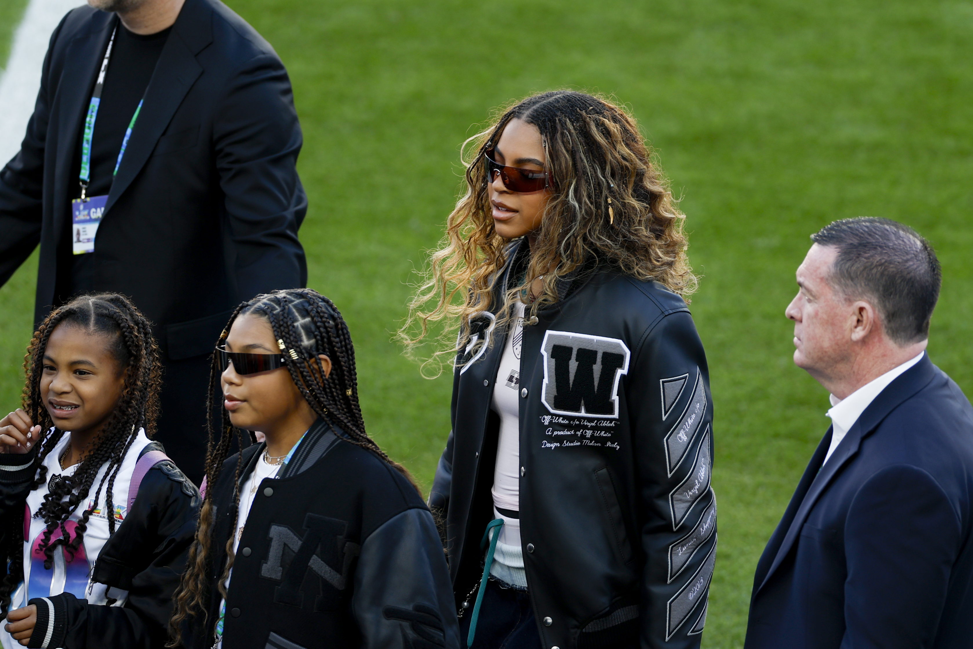 Blue Ivy walks across the field during pregame with her younger sister Rumi at her side, surrounded by family and staff. The sisters move calmly through the scene as attention follows them from the sidelines.