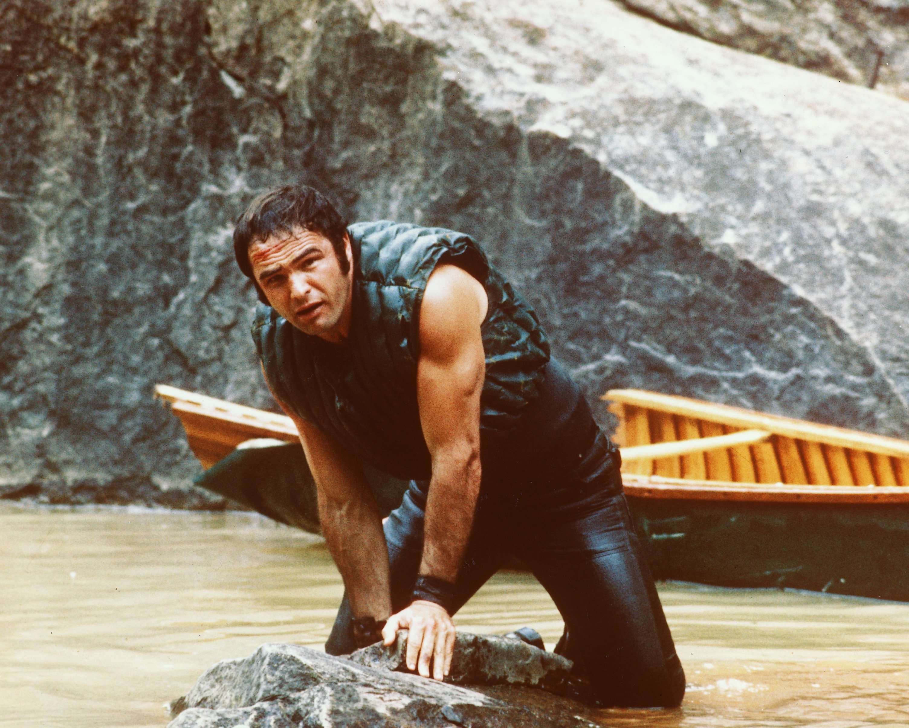 , holding onto some rocks as he struggles in the water with his boat behind him in a publicity still issued for the film, "Deliverance," in 1972. | Source: Getty Images
