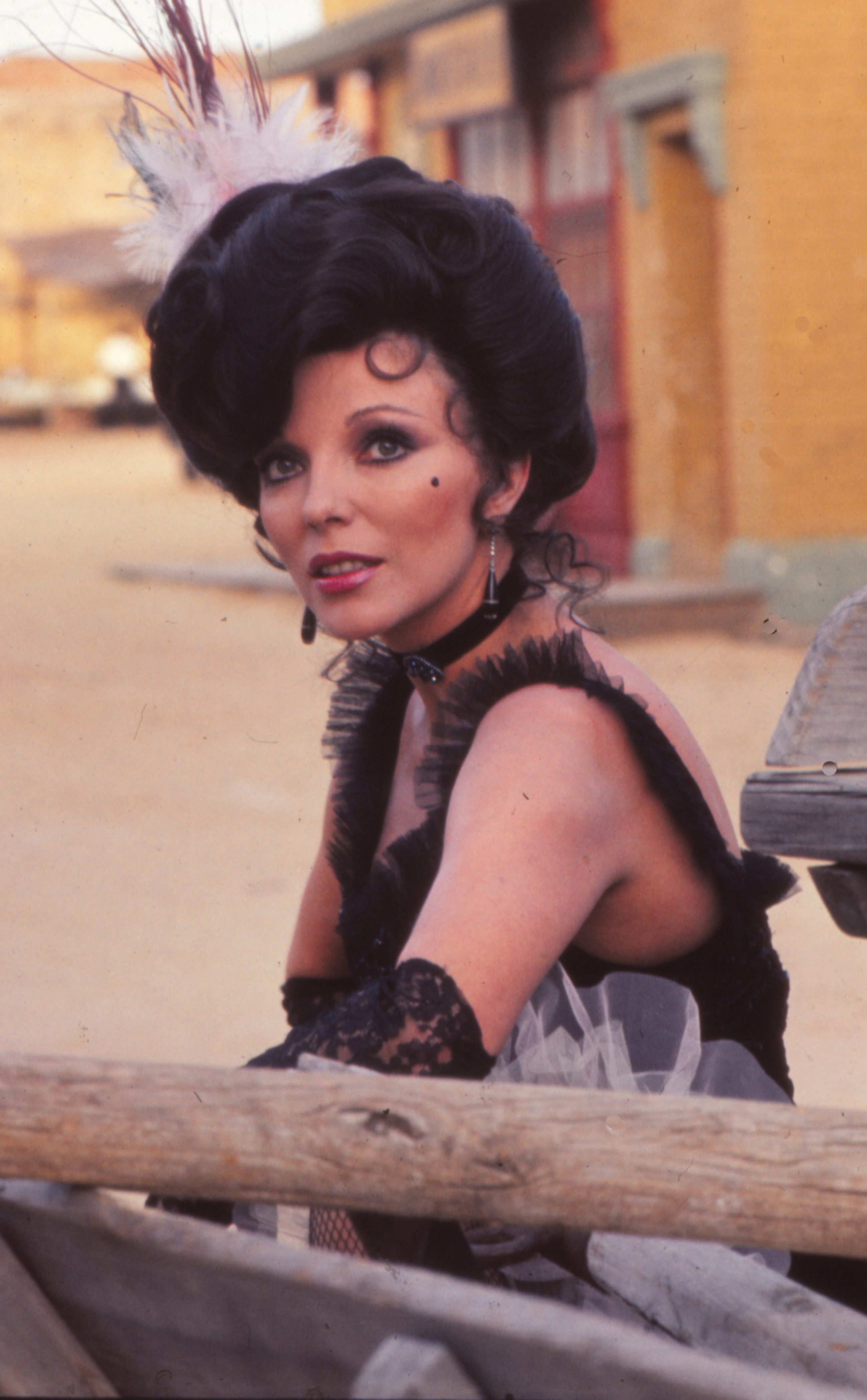 Joan Collins is seen on the set of "La llamada del lobo" in Madrid, Spain, dressed in period costume. She wears a black lace corset dress with tulle and feathered accents, long lace gloves, and a black choker. Her dramatic updo is adorned with a feathered headpiece, and she finishes the look with bold eyeliner and a beauty mark on her cheek.
