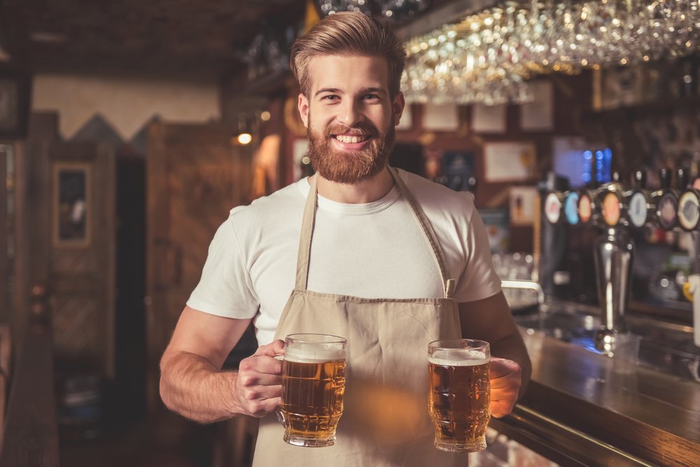 Ein Barkeeper in Schürze, der Bier in der Hand hält, während er in einer Kneipe in der Nähe des Tresens steht. | Quelle: Shutterstock