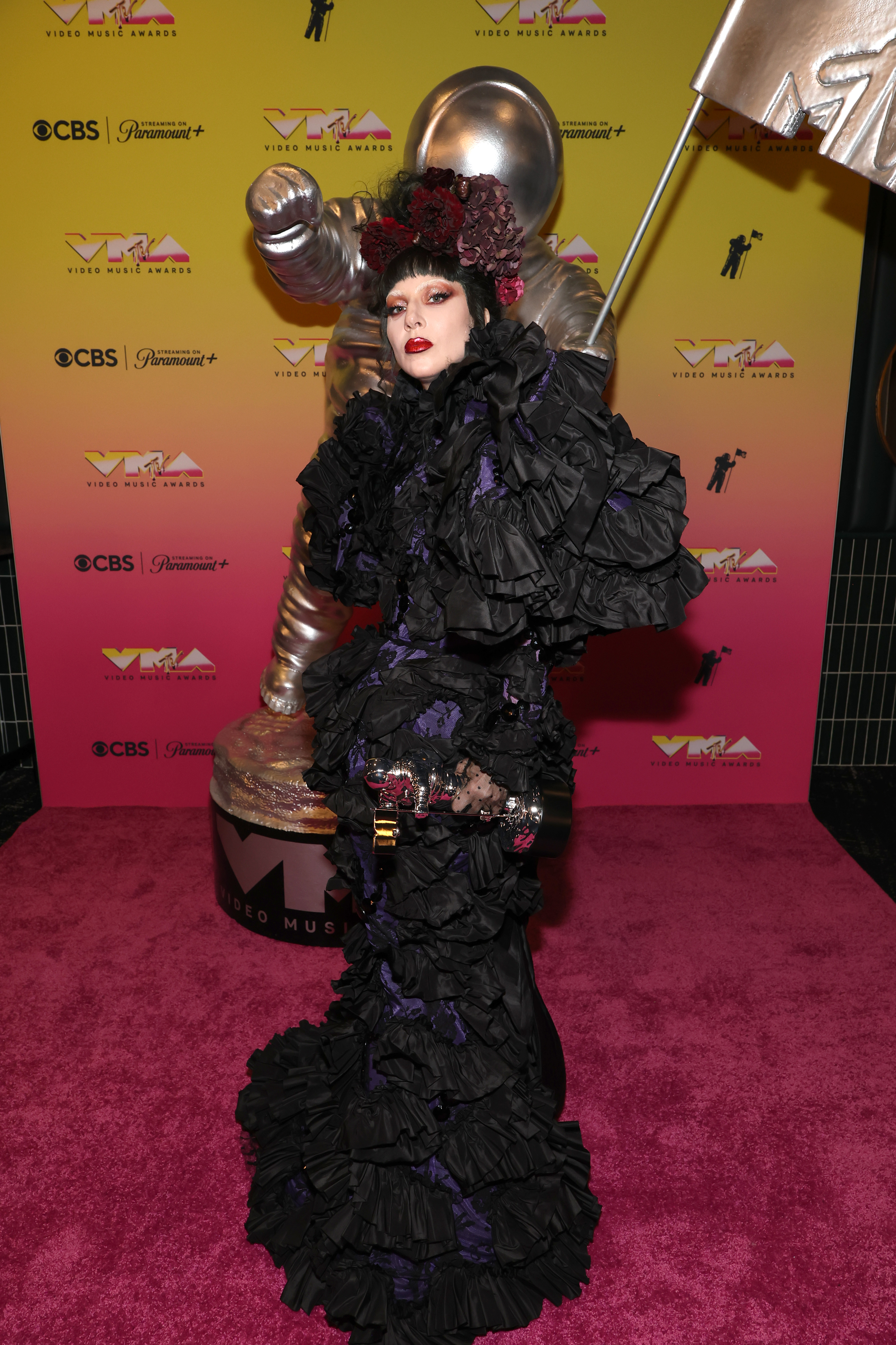 Lady Gaga poses with her Artist of the Year award, wearing a dramatic, ruffled black gown with purple accents and an eye-catching floral headpiece.