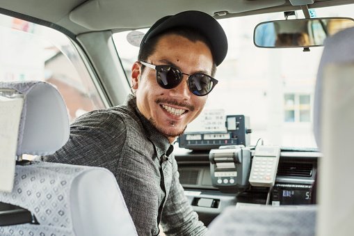 A man wearing sunglasses and baseball hat in the passenger seat of a car turning around and smiling | Photo: Getty Images