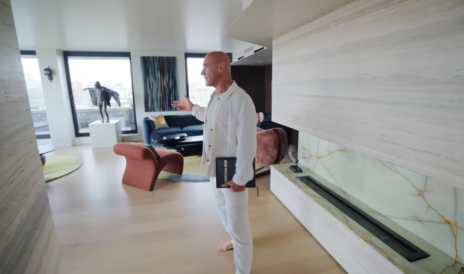 The monumental fireplace creates a boundary without building a conventional wall between the living areas. Its pale stone surface feels almost architectural enough to be a room in itself. | Source: YouTube/Architectural Digest