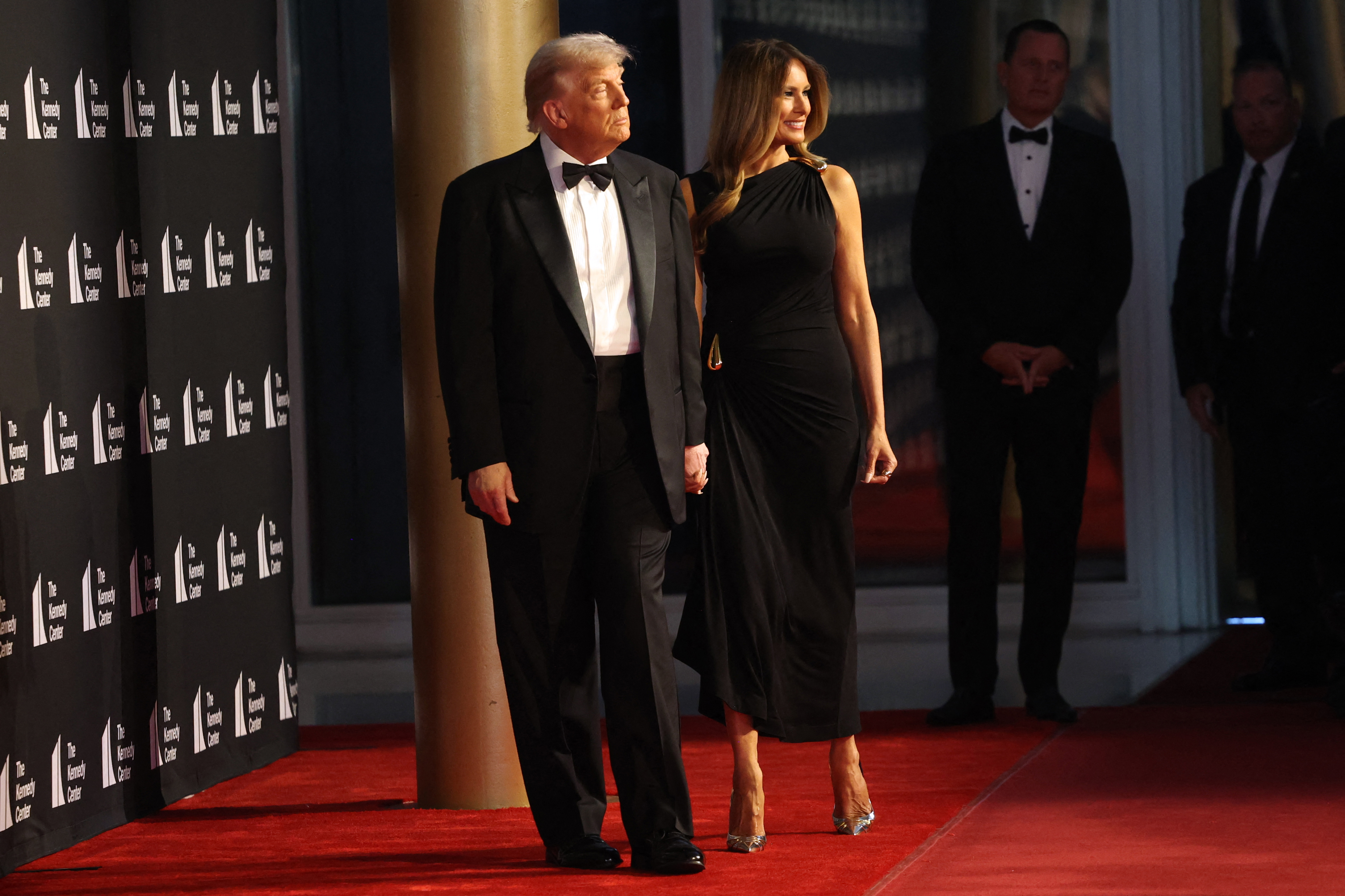 Donald and Melania on the red carpet, both looking toward the photographers. Her gown falls gracefully to mid-calf, paired with metallic heels that catch the light. Melania smiles slightly, while Donald stands composed at her side.