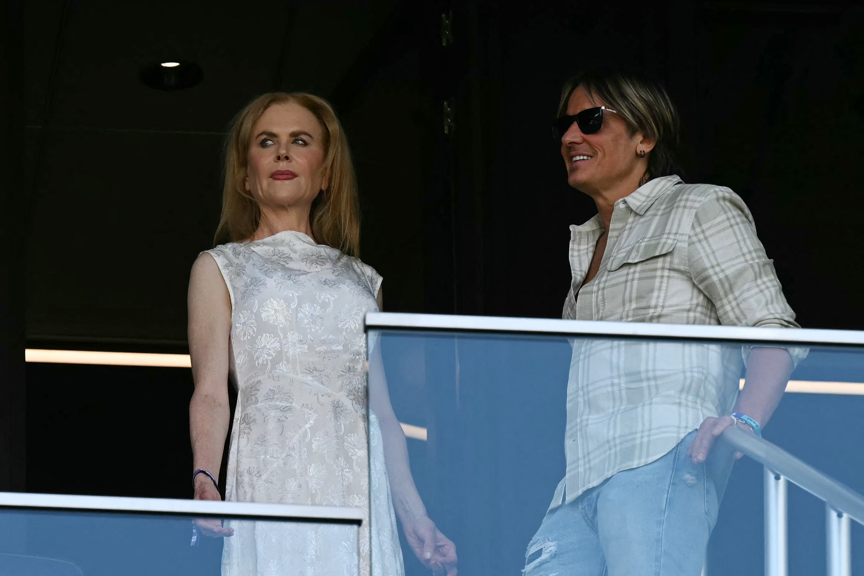 Framed against the shadows of the VIP box, Nicole Kidman stands poised beside Keith Urban, who keeps his cool behind dark shades as they take in the match from above.