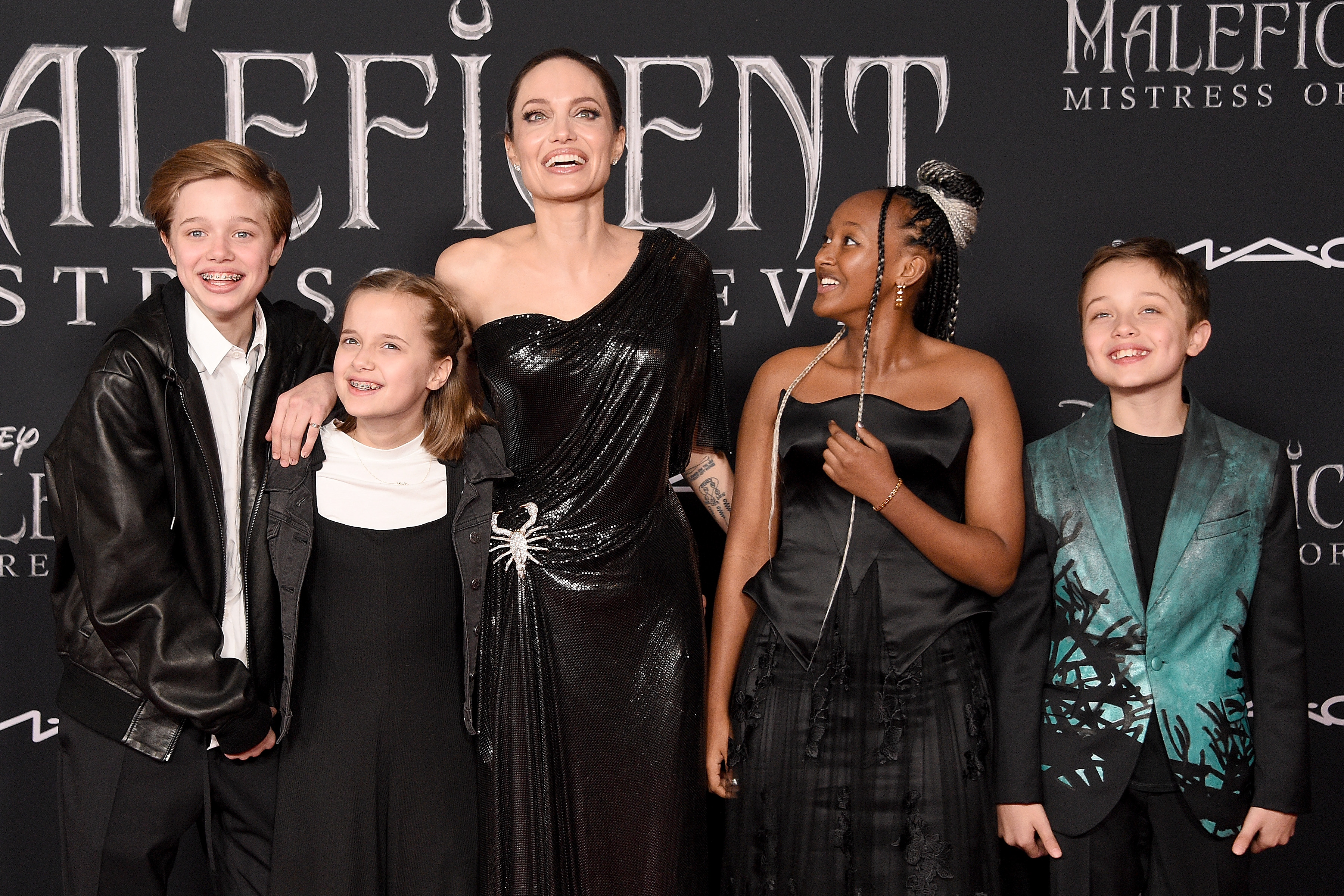 Shiloh Jolie-Pitt smiles on the red carpet at the "Maleficent: Mistress of Evil" premiere alongside Angelina Jolie and her siblings. In a black jacket layered over a white shirt, her look feels more relaxed yet refined. The moment marks a clear step toward a more polished, evolving style.