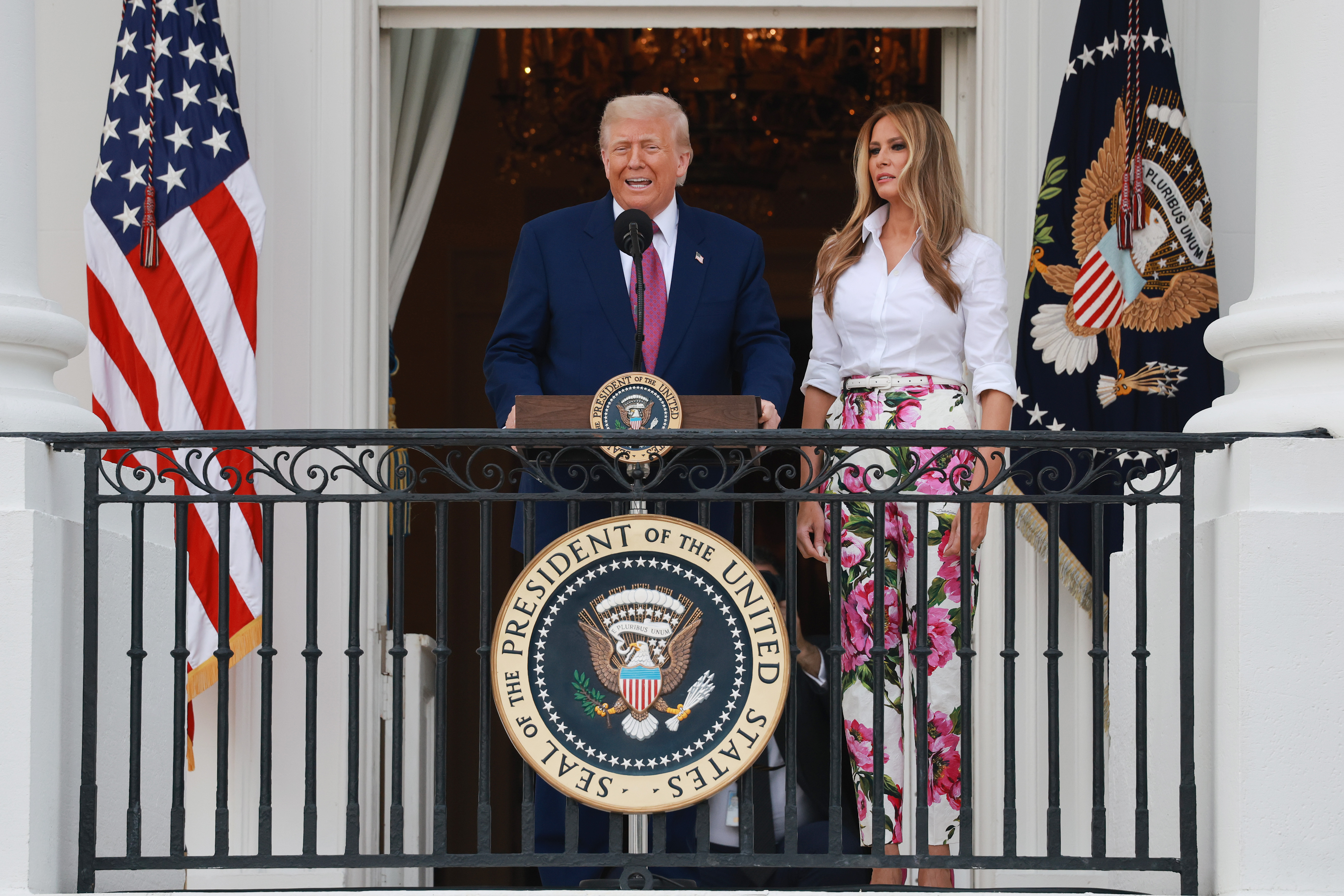 Framed by American and presidential flags, Donald Trump addresses the crowd while Melania stands quietly beside him on the White House balcony. Their positioning and expressions reflect a moment of formality and public presence.