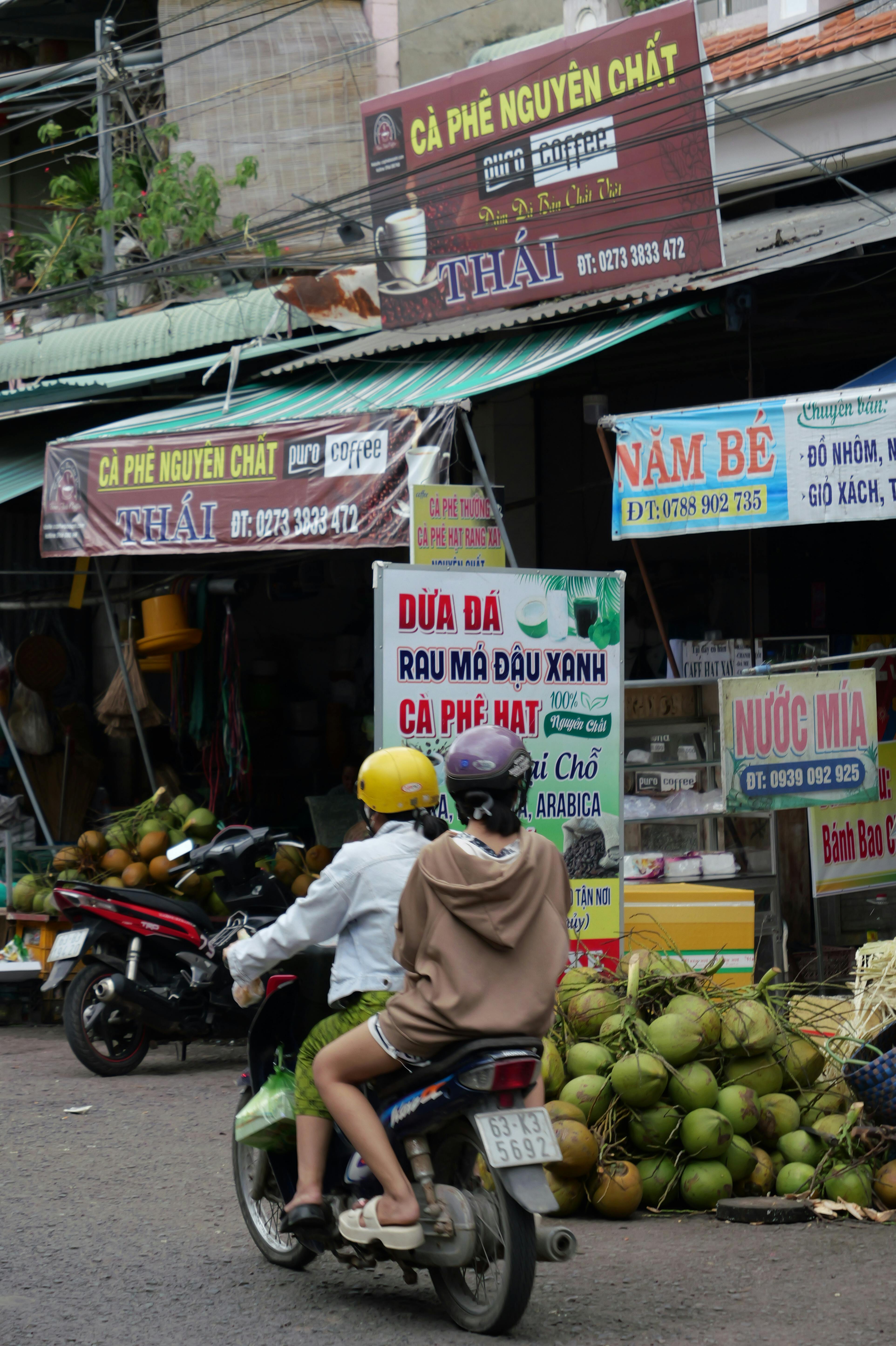 Two riders pass market stalls and street signs in a busy Vietnamese neighbourhood filled with local shops and produce | Source: Pexels