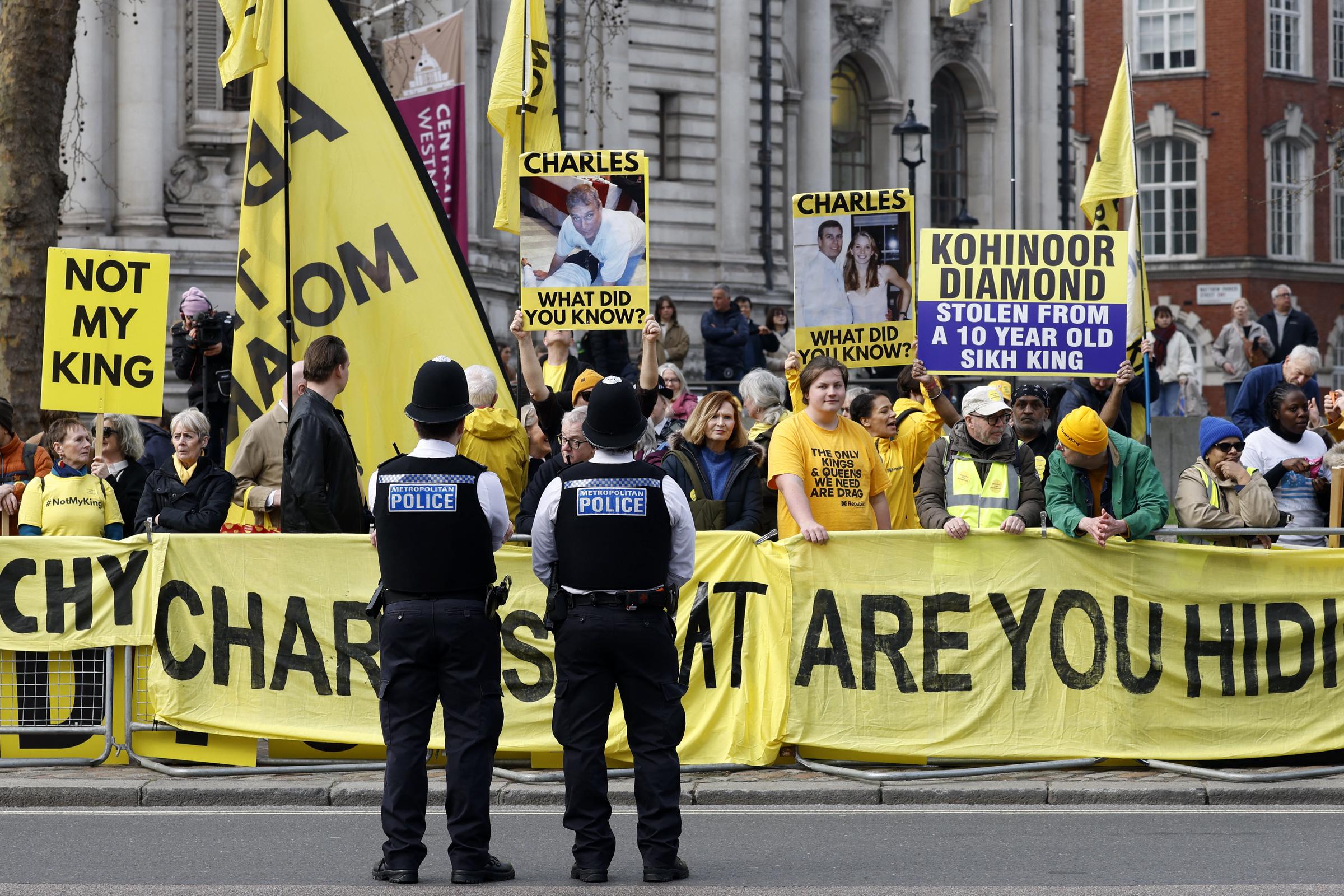 Two Metropolitan Police officers stood with their backs to the camera facing a crowd of anti-monarchist protestors behind bright yellow barriers outside Westminster Abbey on 9 March 2026, the demonstrators brandishing "Not My King" placards, photos of King Charles III, and a large banner reading "Kohinoor Diamond Stolen From A 10 Year Old Sikh King."