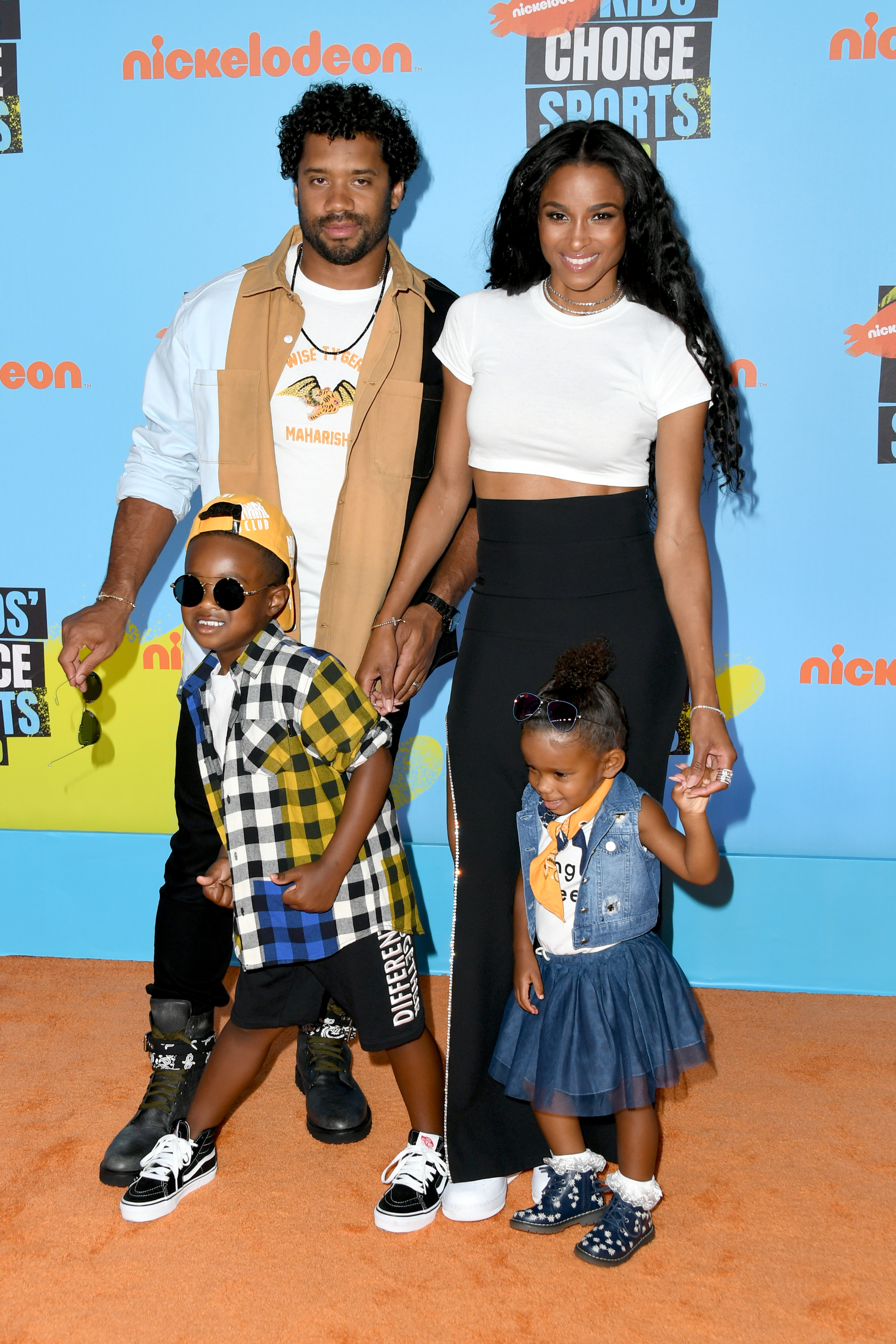 Ciara and Russell Wilson pose with their children, Future Zahir Wilburn and Sienna Princess Wilson, standing close together as they hold hands and present a united family front. All are dressed stylishly, with their smiles and relaxed posture creating a warm and cheerful atmosphere.