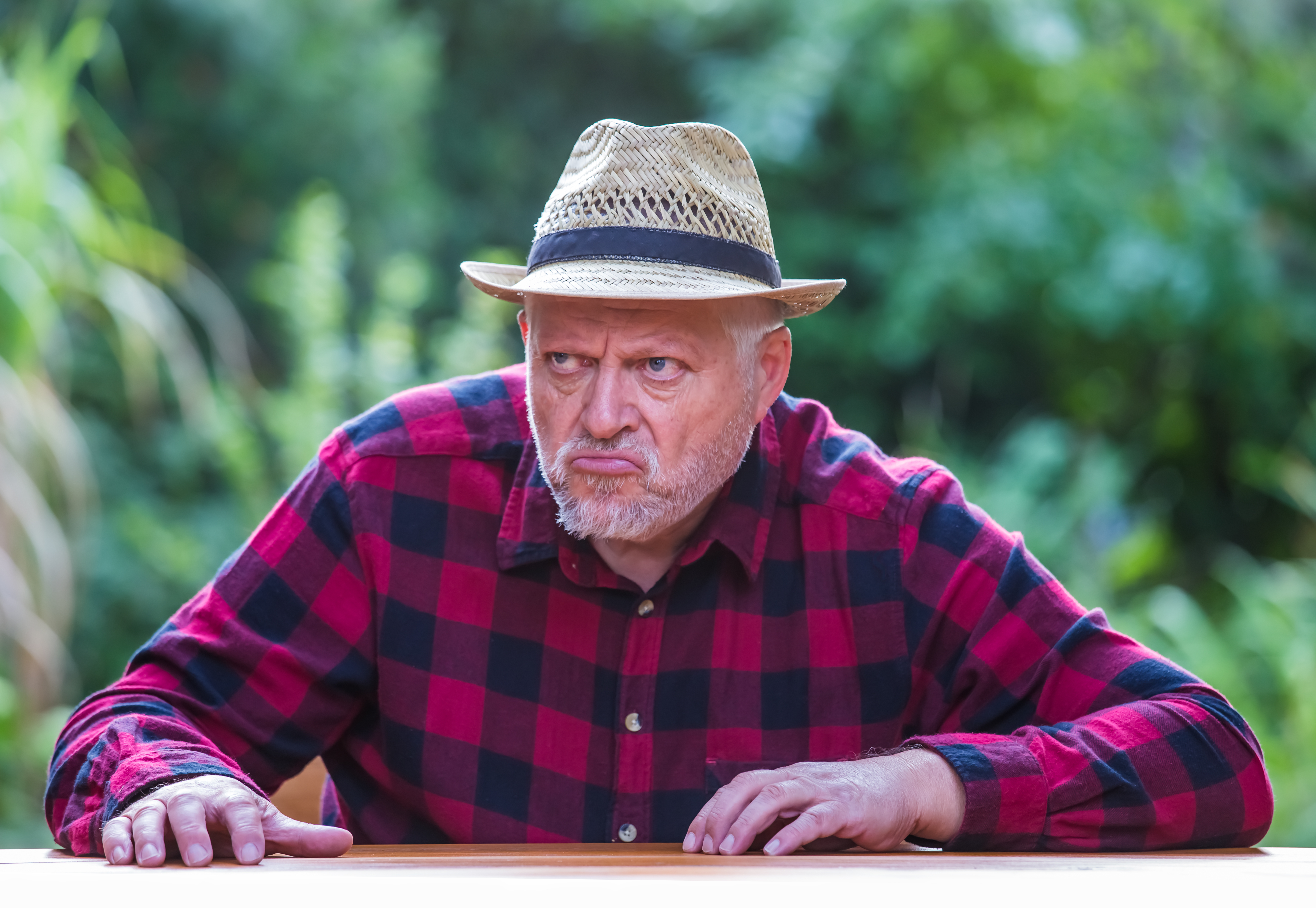 An elderly man in a hat is sitting with bad mood at a table in the garden. | Source: Shutterstock