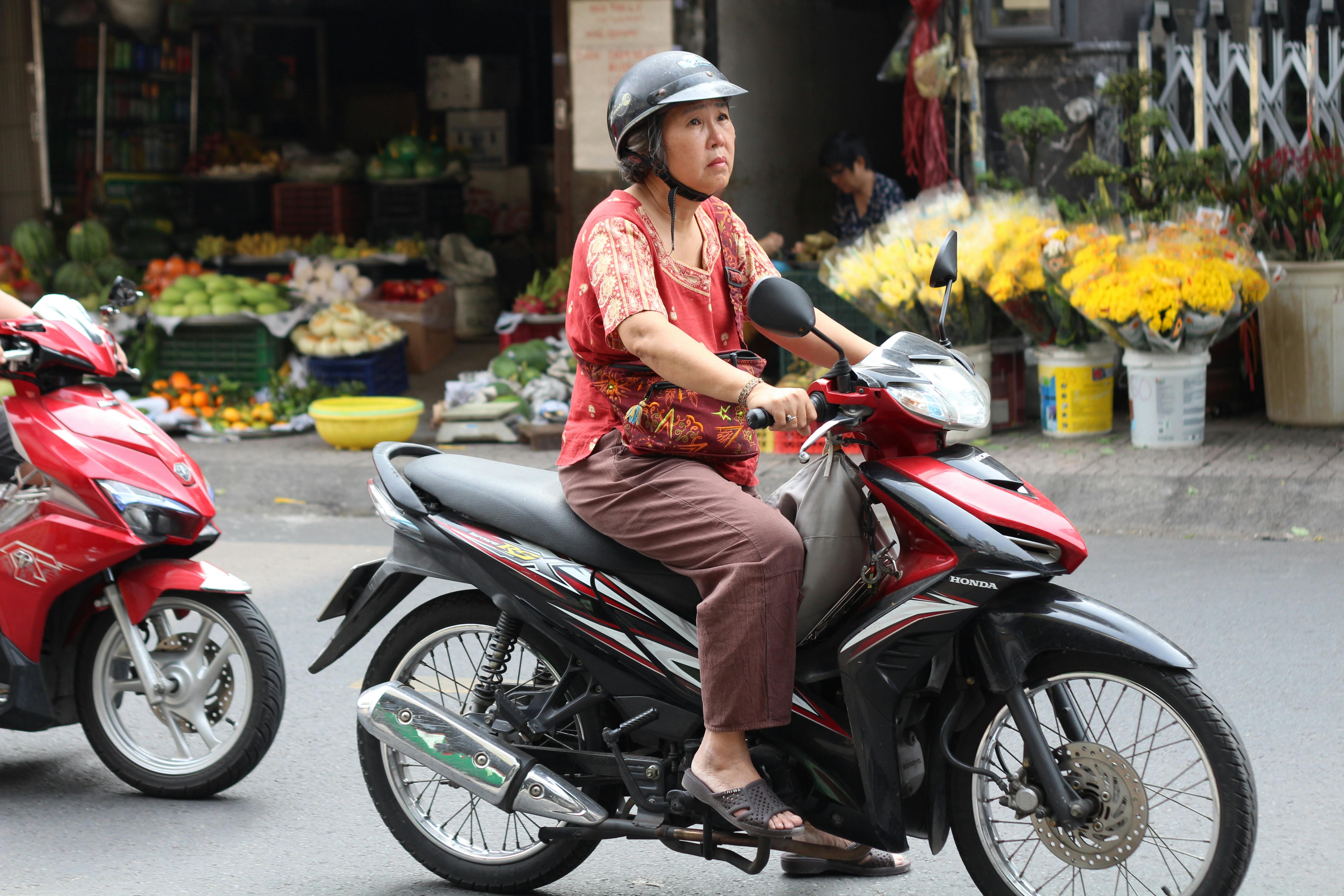 A woman rides a motorbike through a busy Vietnamese street lined with market stalls and flowers | Source: Pexels